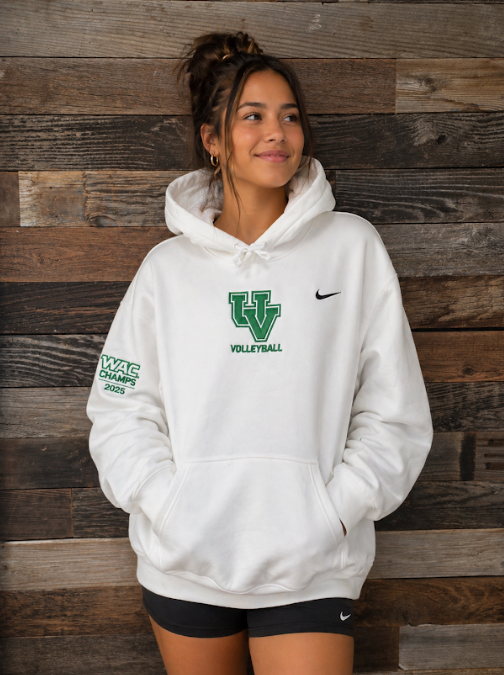 Woman in white hoodie and black shorts standing against a wooden wall, looking to the side.