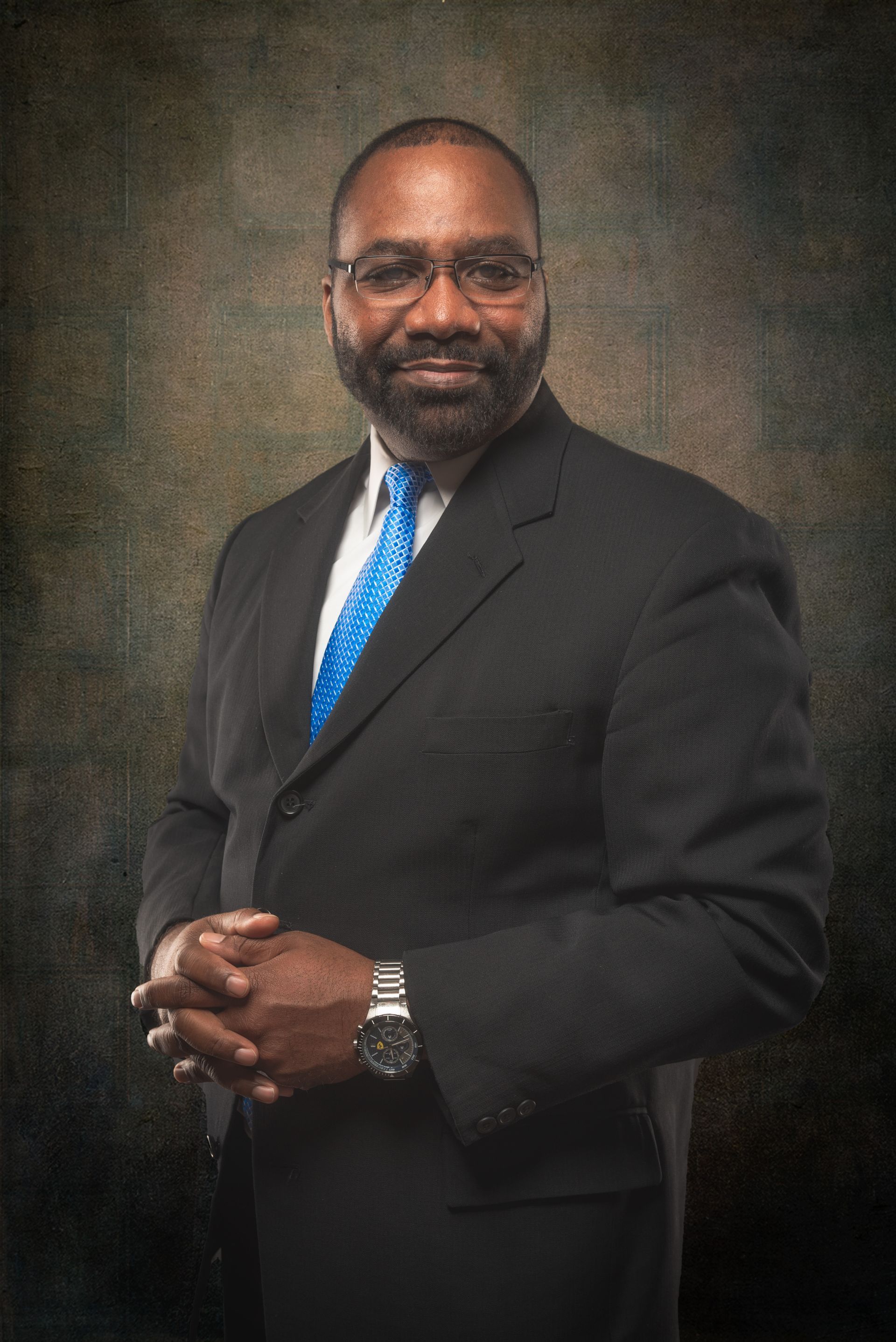 Man in a suit, hands clasped, smiling at camera against a mottled dark background.