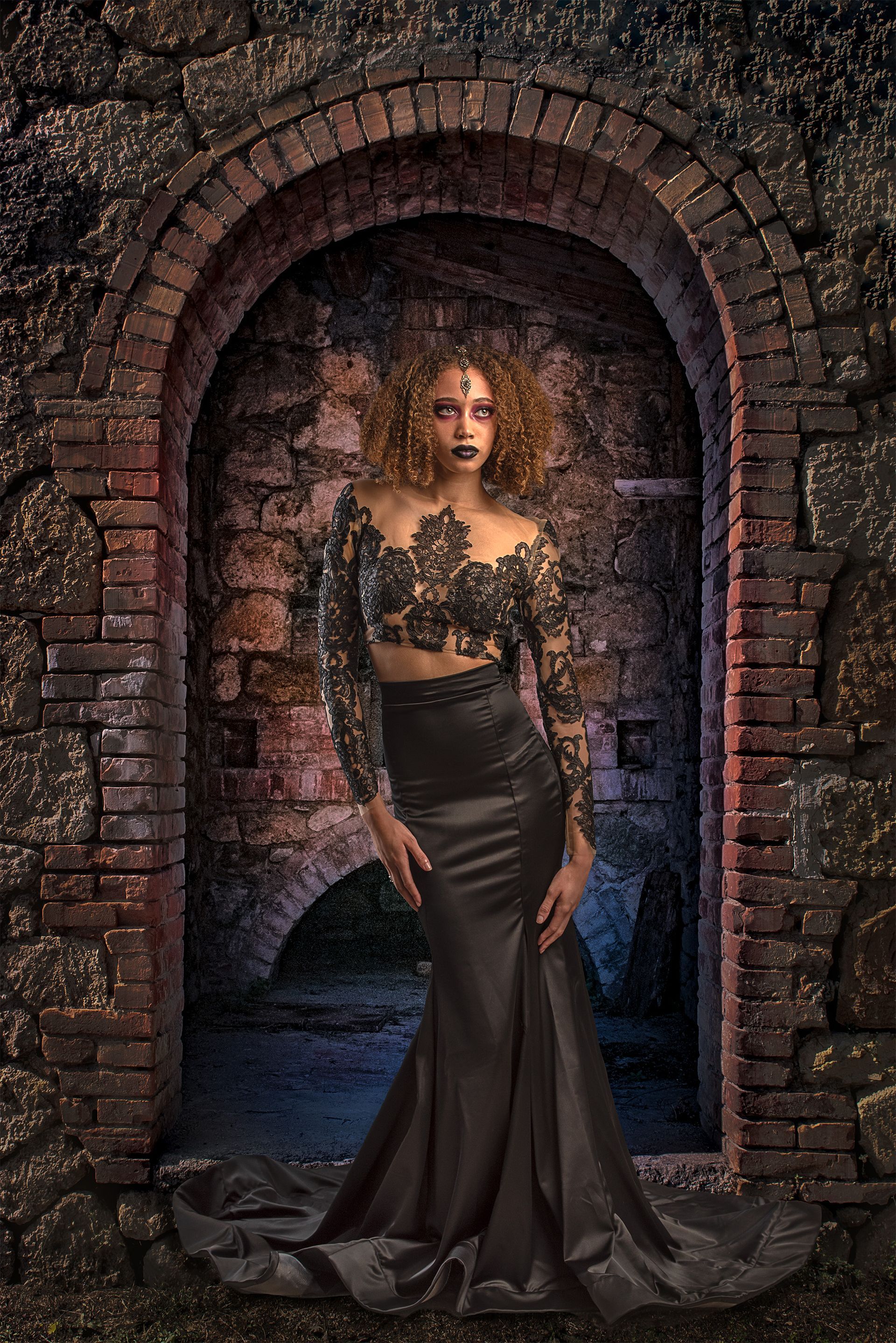 Woman in black lace top and satin skirt, standing in arched brick doorway.