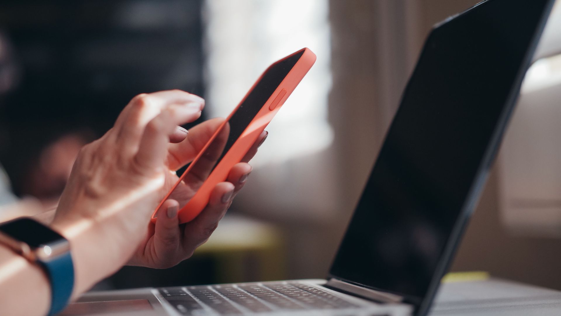 Hands holding an orange smartphone beside an open laptop on a desk
