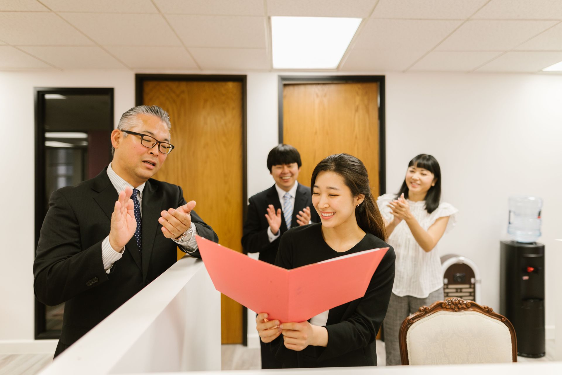 Four coworkers in an office applaud as a woman holds a red folder and smiles