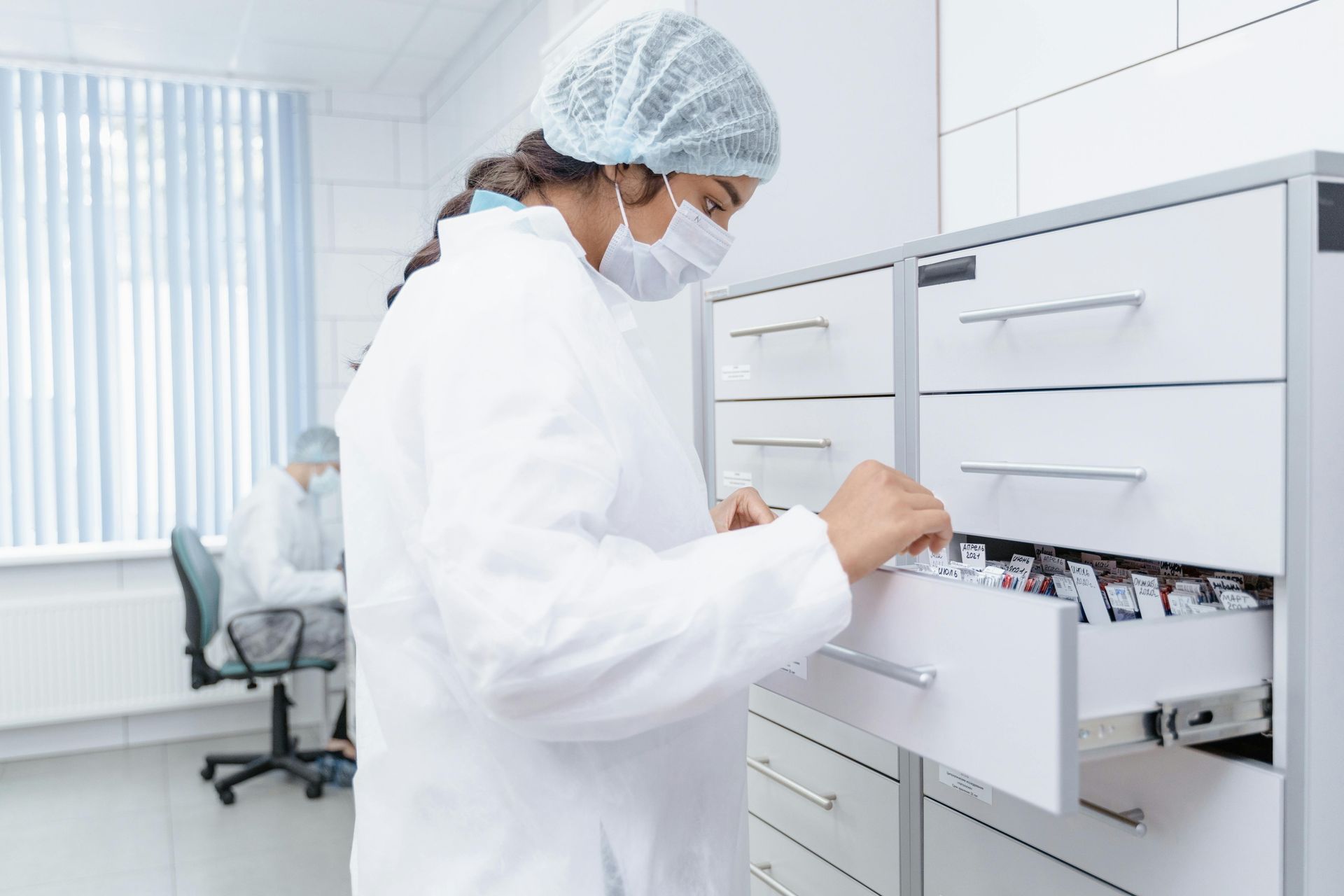 A woman in a lab coat and mask is looking in a drawer.