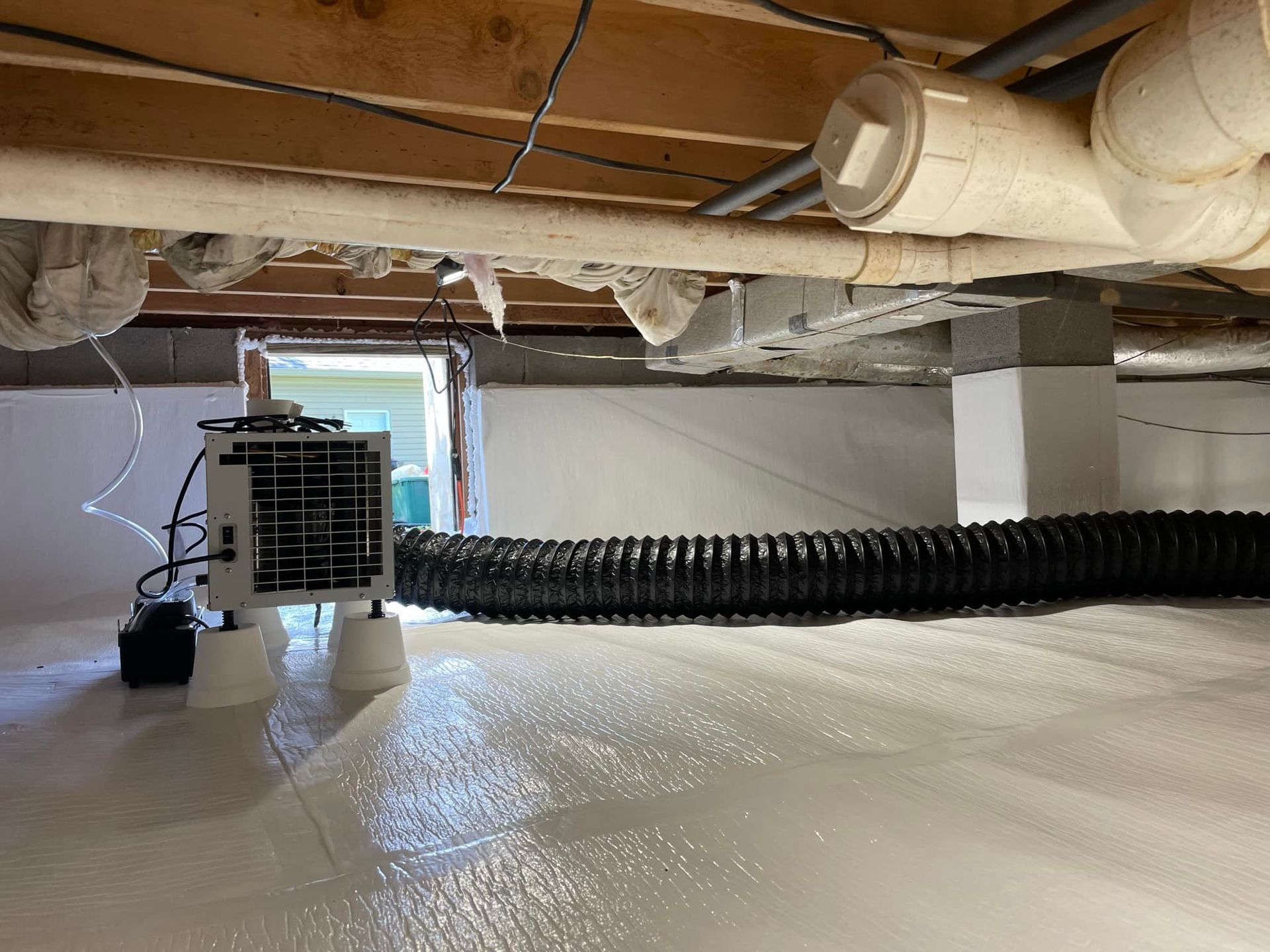 A crawl space with a dehumidifier and black hose on white vapor barrier under a house.