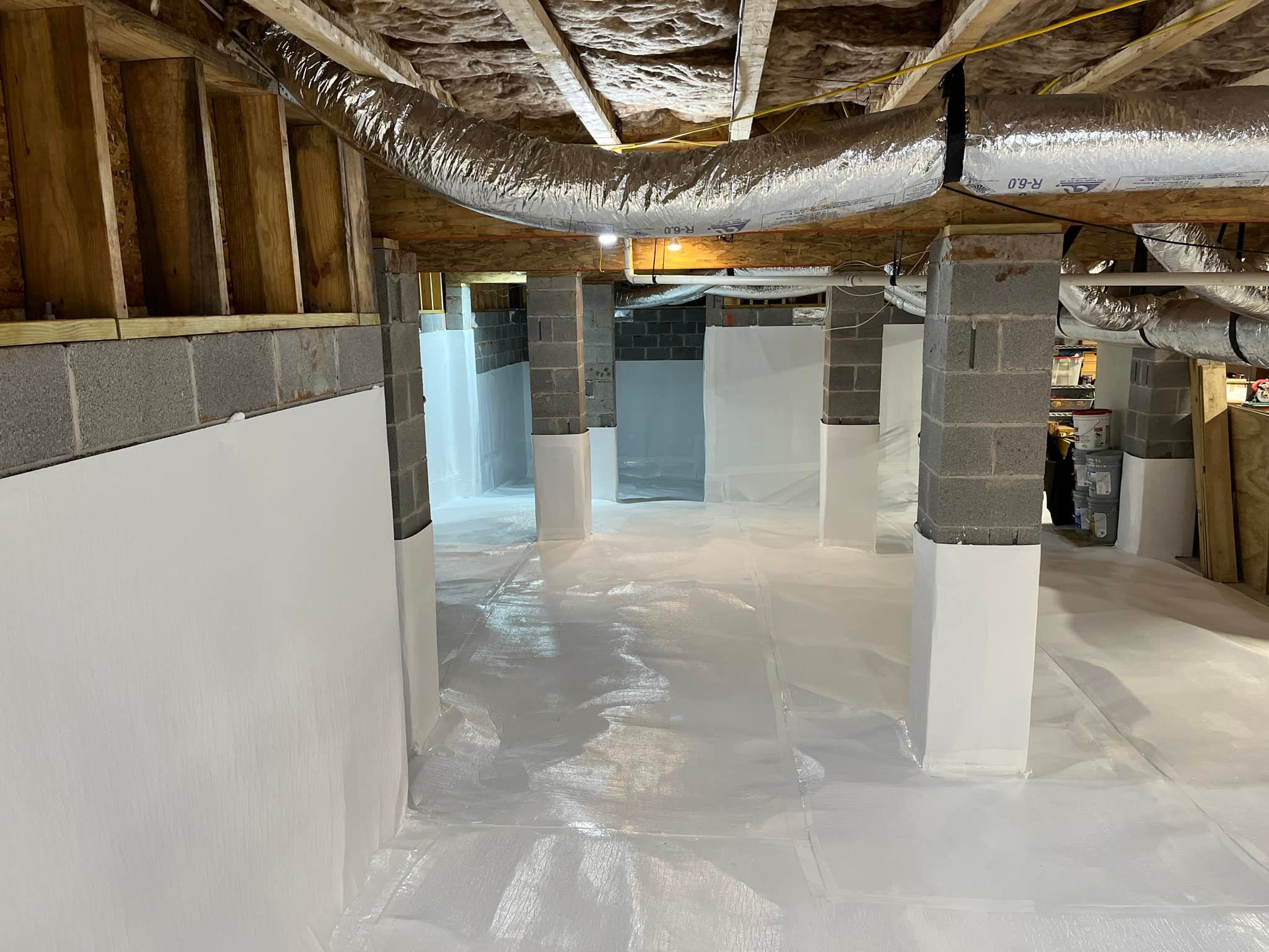 A crawlspace with white walls and floor covering, gray support columns, and wooden beams above.