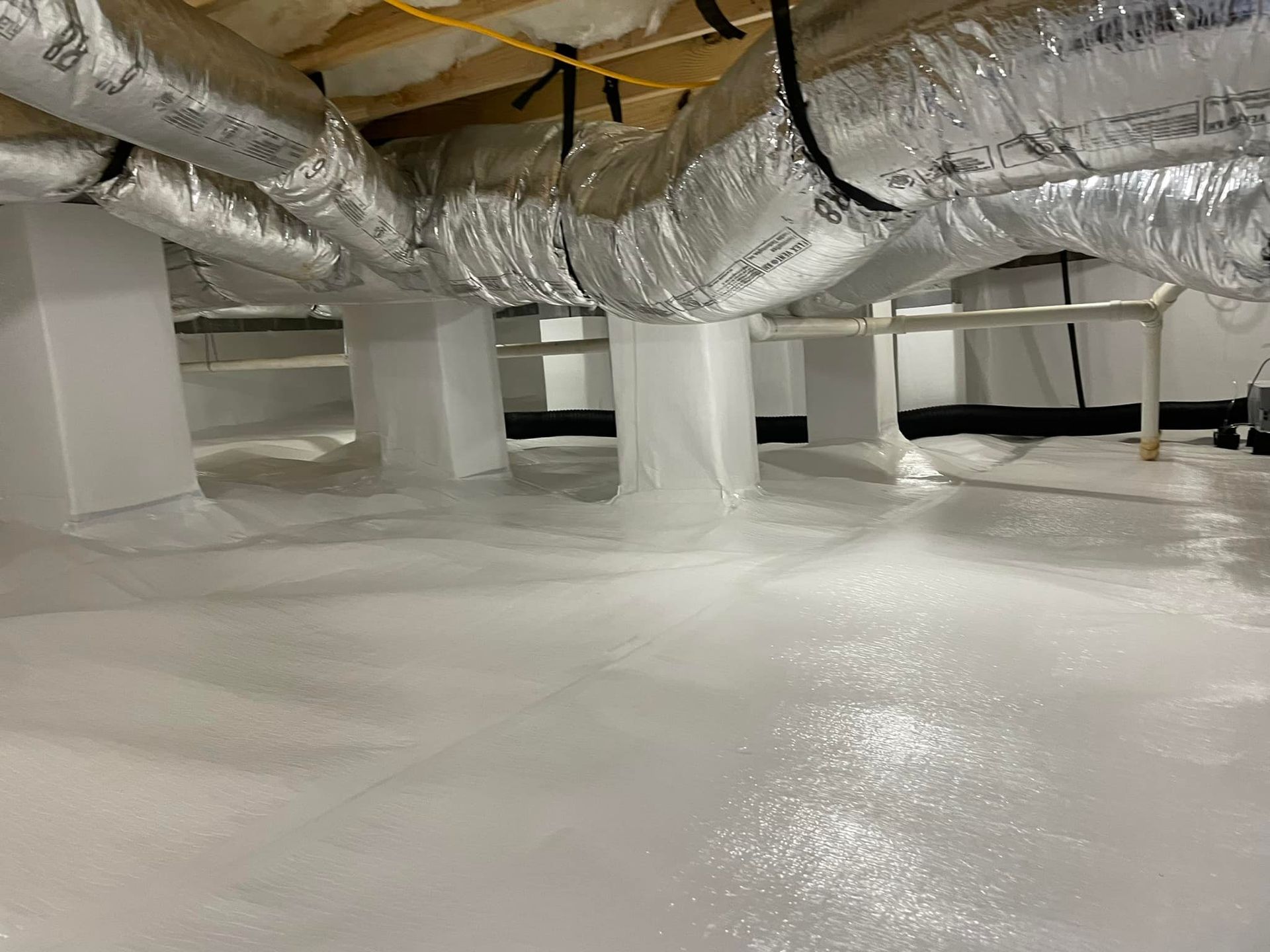 White crawlspace with vapor barrier on floor, insulation on pillars, and ductwork overhead.
