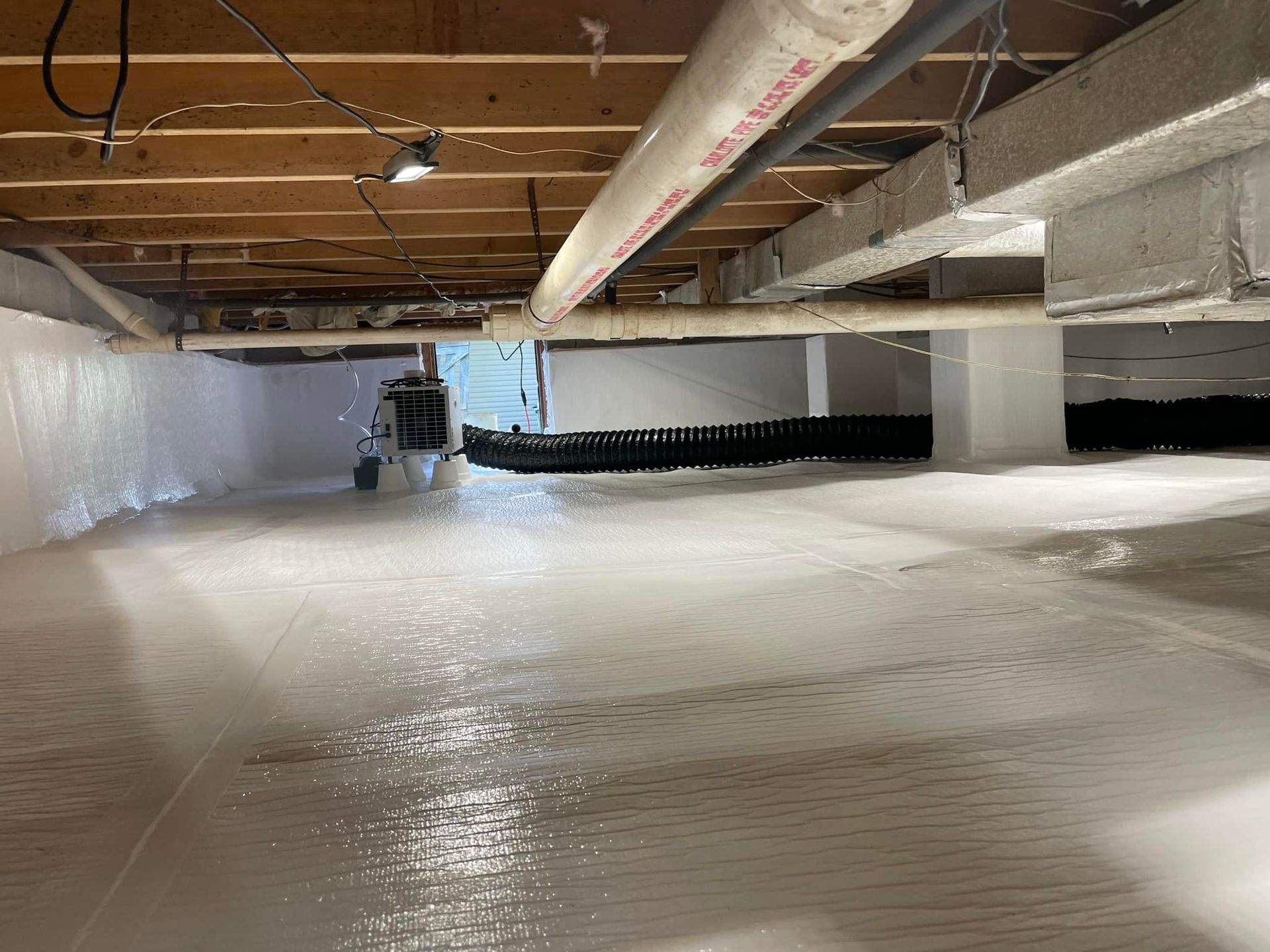 Crawl space with white vapor barrier on walls and floor, plumbing, and ductwork overhead.