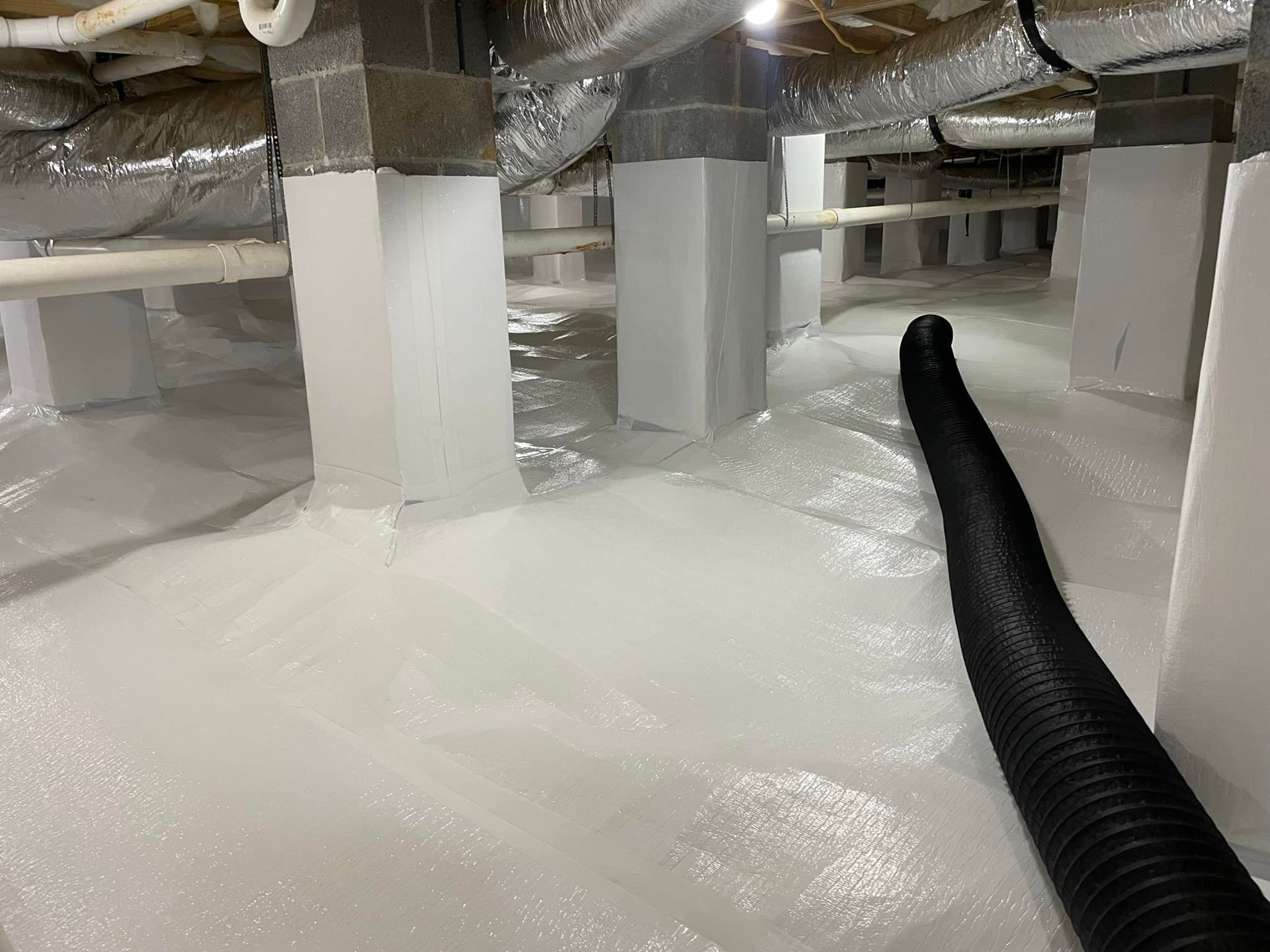 White crawlspace with vapor barrier on the floor, supporting beams, and ductwork. Black hose extends into space.