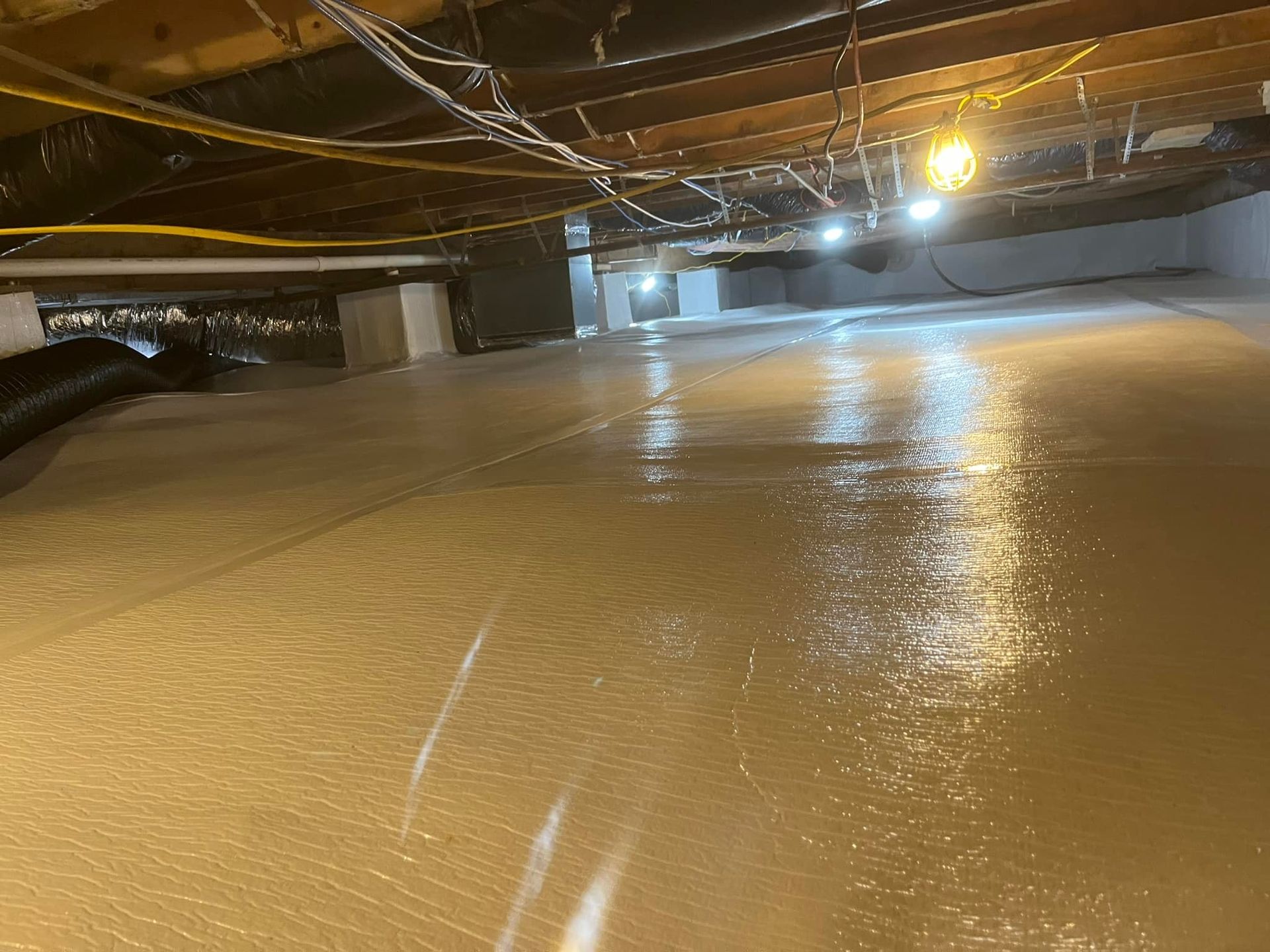 A crawl space with insulation on the ground and overhead beams. It's brightly lit.