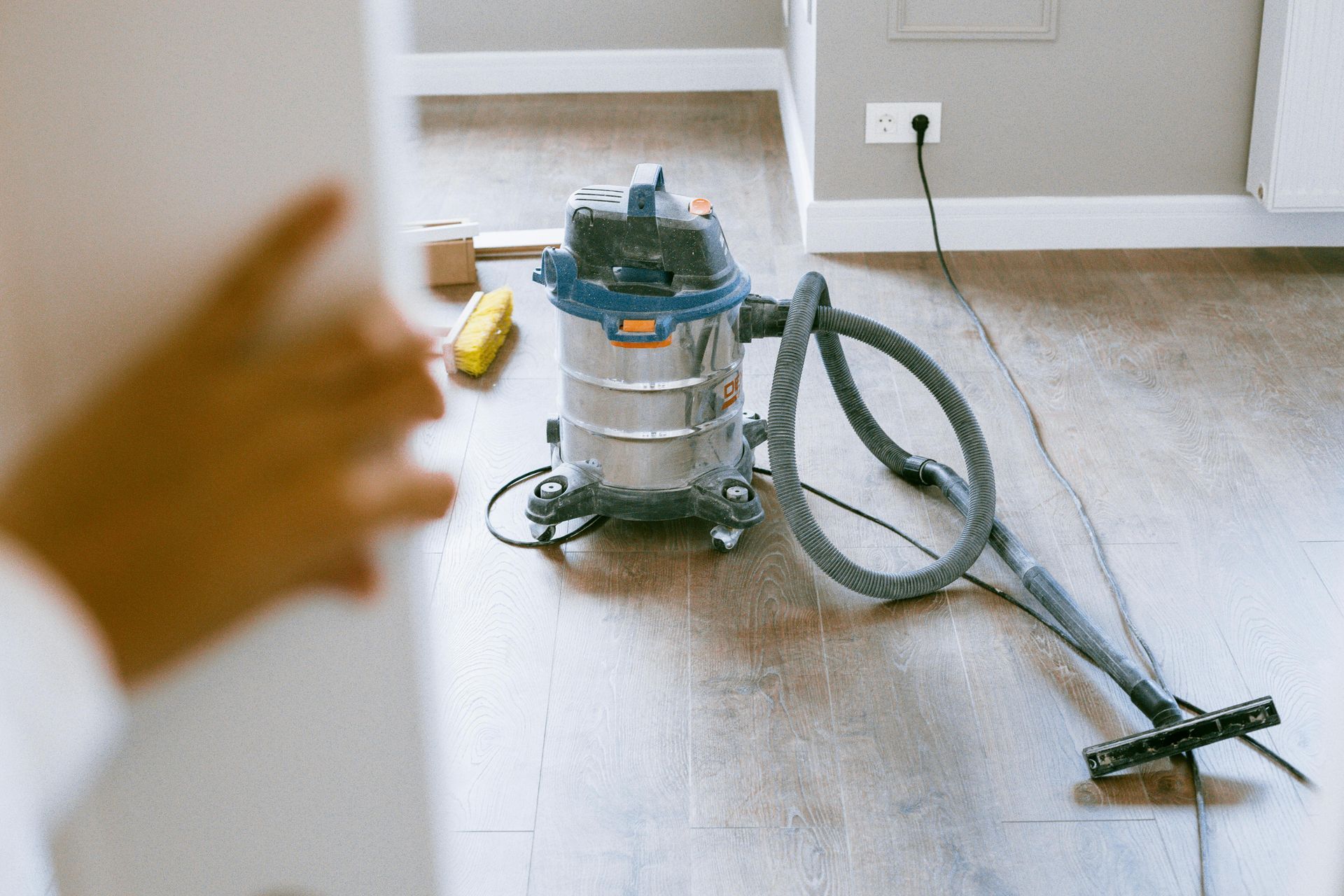 A wet/dry vacuum cleaner sits on a wooden floor near a wall with an outlet.