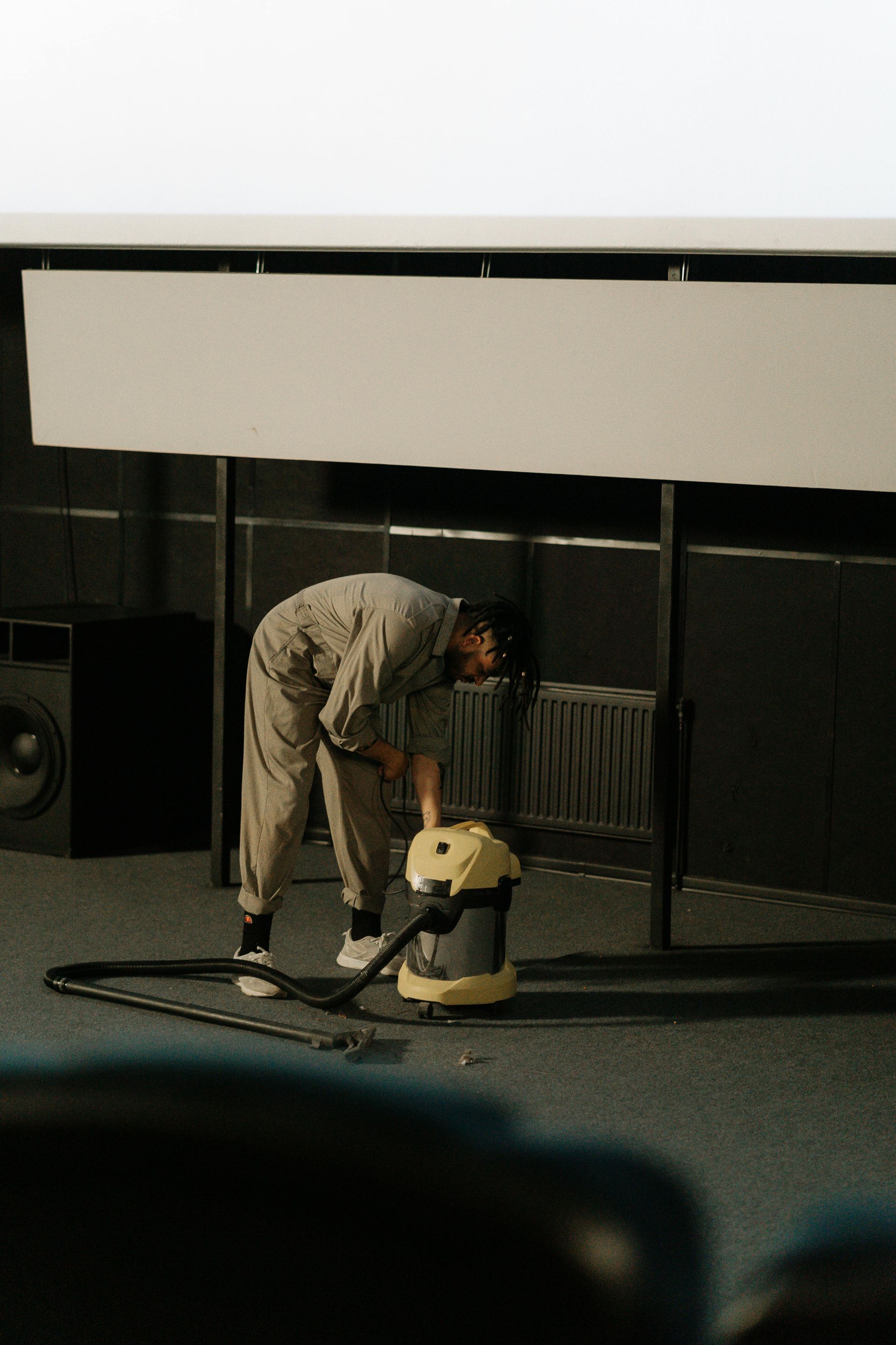 Person in jumpsuit vacuums a dark room with a large screen, near a speaker and metal panel.