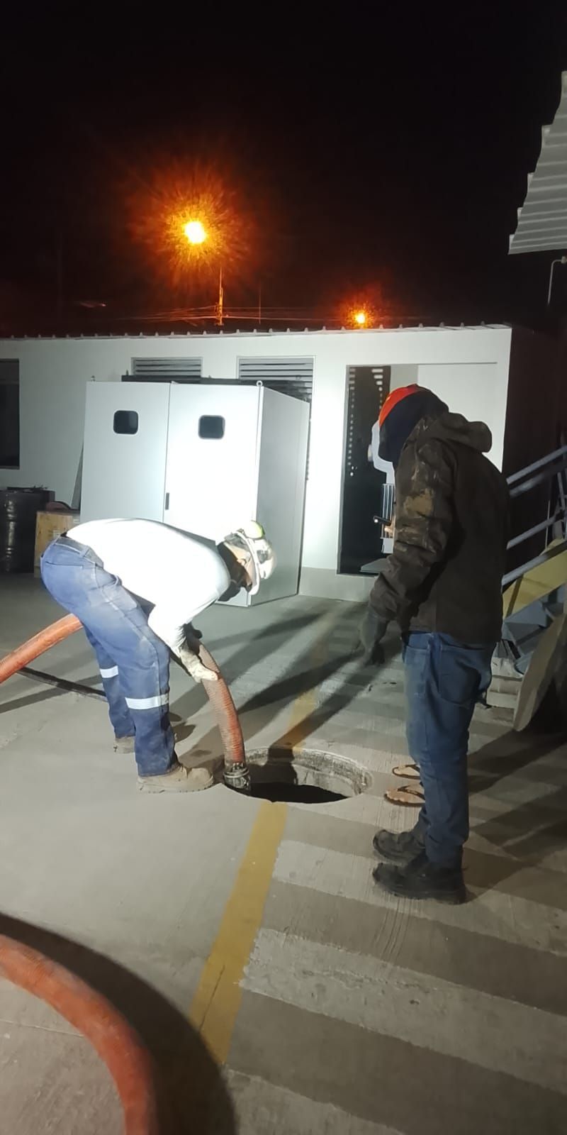 Dos personas trabajan cerca de una alcantarilla por la noche. Una sostiene una manguera, la otra está cerca.