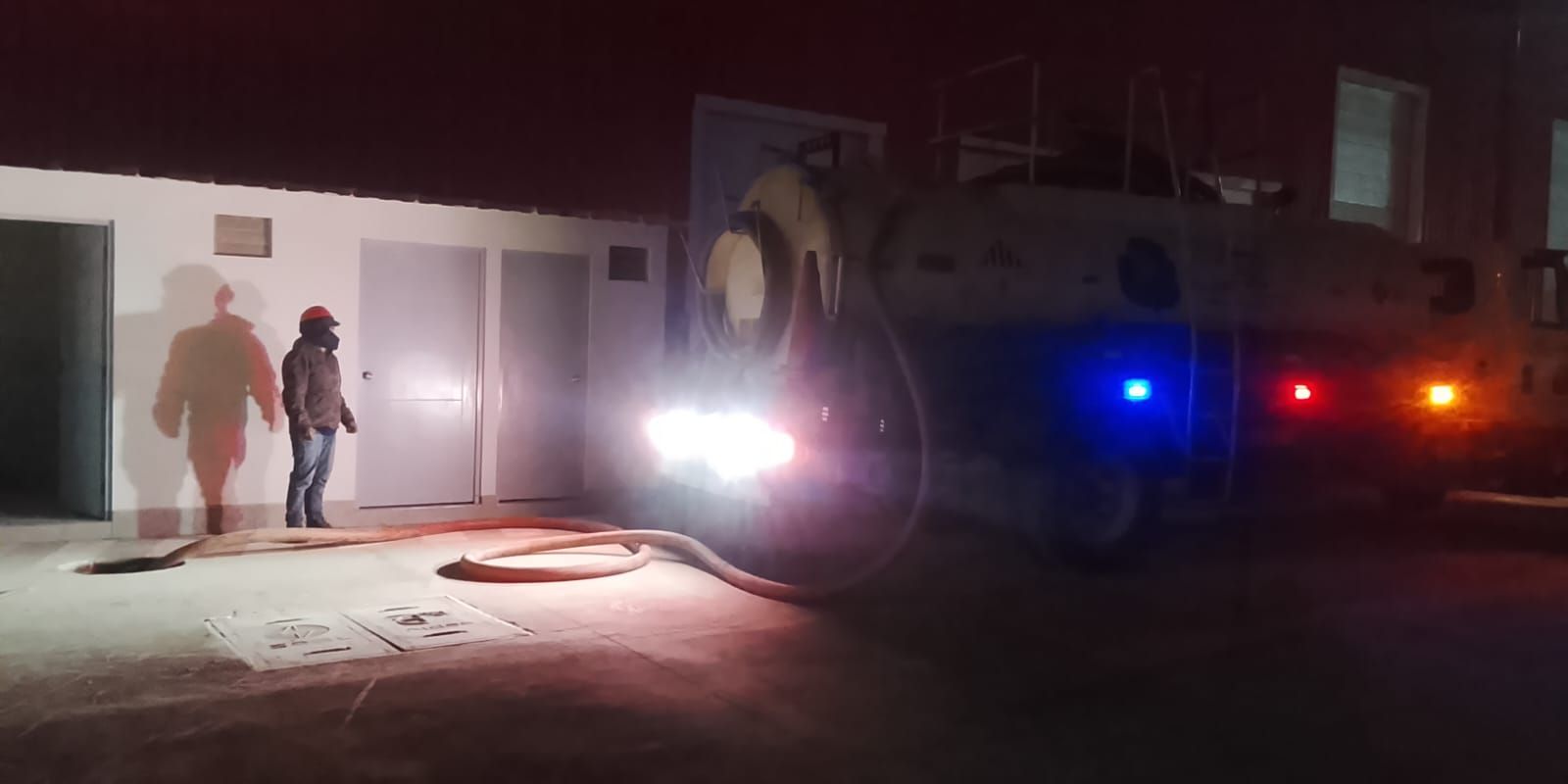 Dos personas están de pie frente a un edificio de noche. Un camión grande está estacionado cerca, con las luces encendidas.