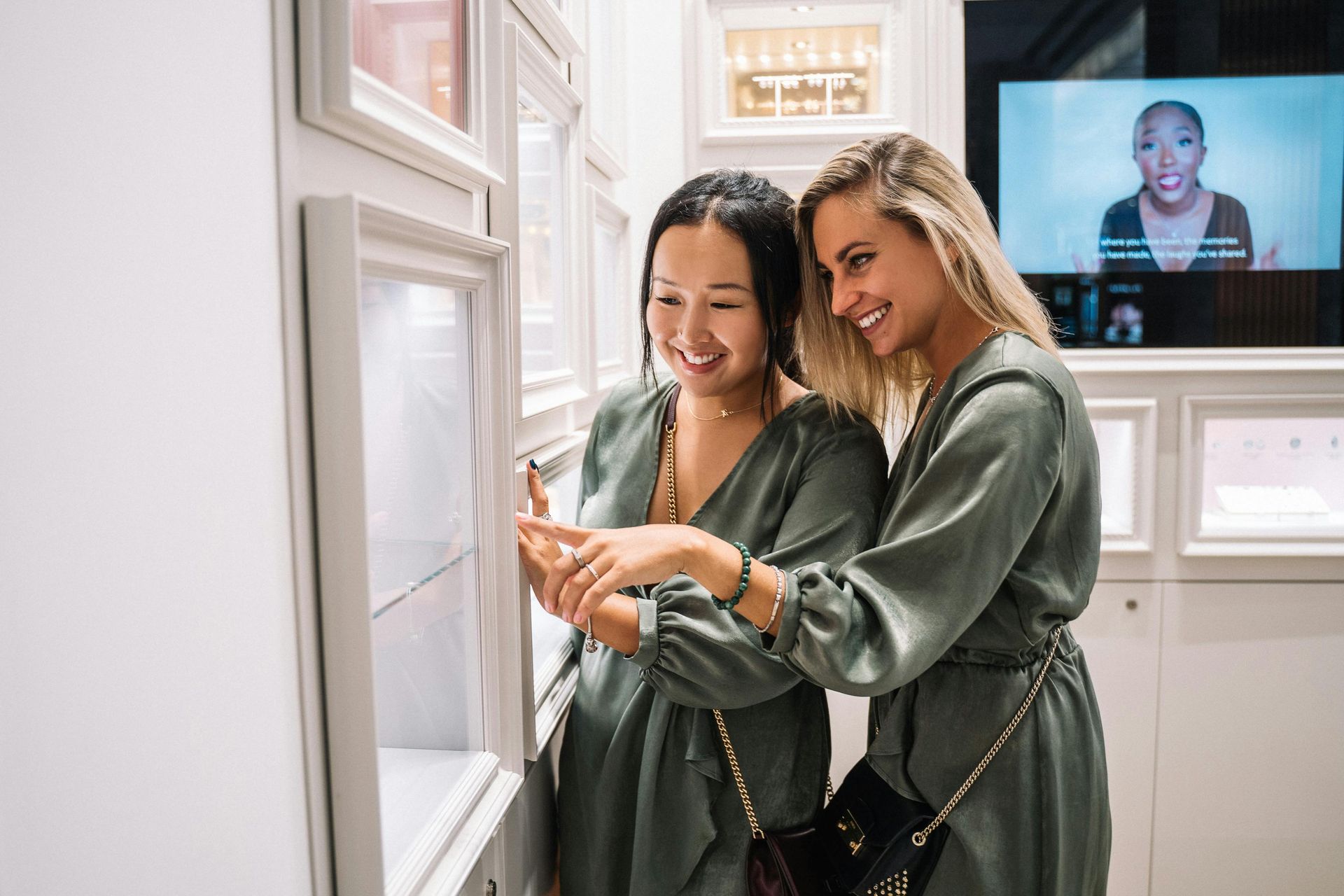 Two women in matching green dresses smiling, looking at a display case in a store.