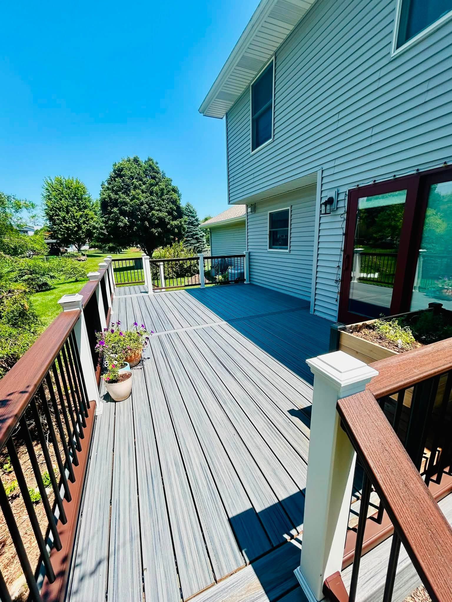 White house deck with railing, potted plants, and a view of trees and lawn on a sunny day