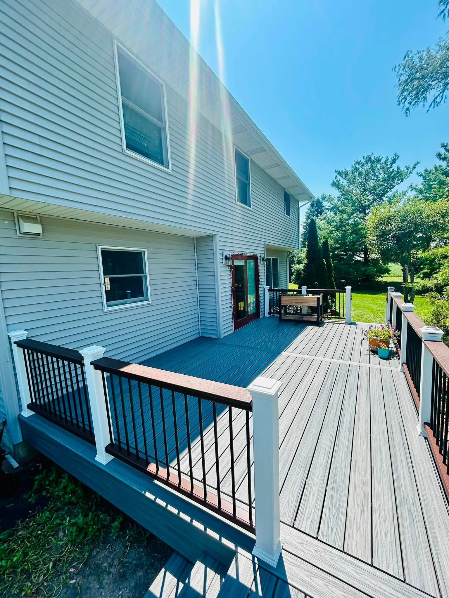 Wooden backyard deck beside a white house, with black railings and outdoor seating in bright sunlight.