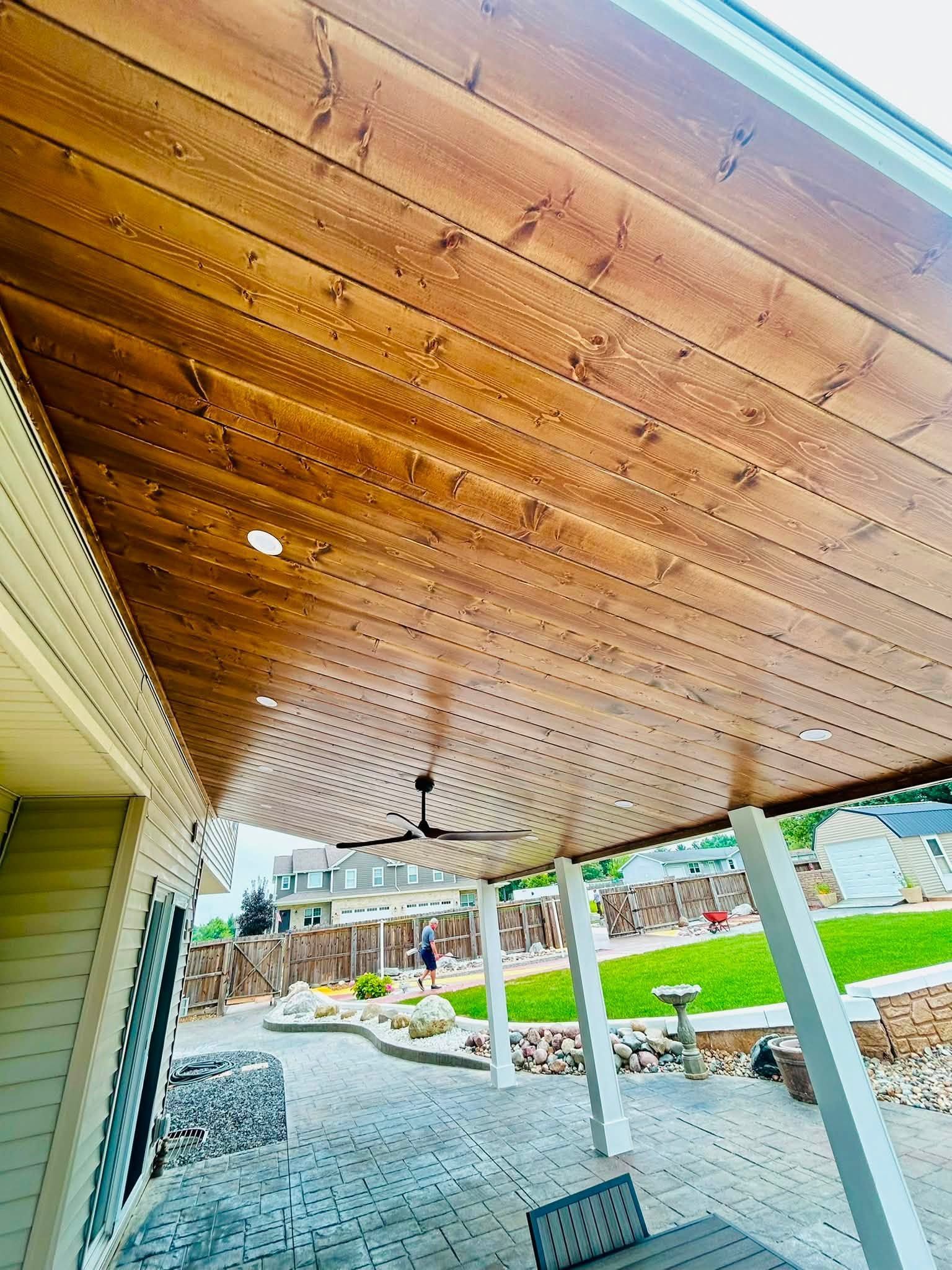 Covered patio with wood-paneled ceiling, string lights, and white columns overlooking a landscaped yard