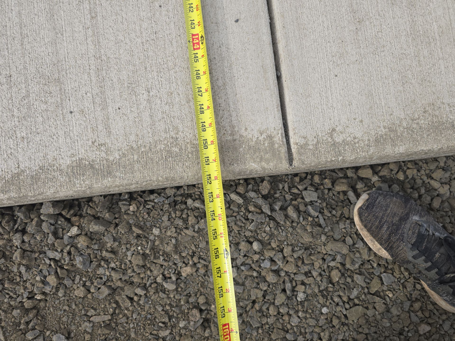 Measuring tape standing beside a concrete wall and gravel ground, with a shoe at right edge