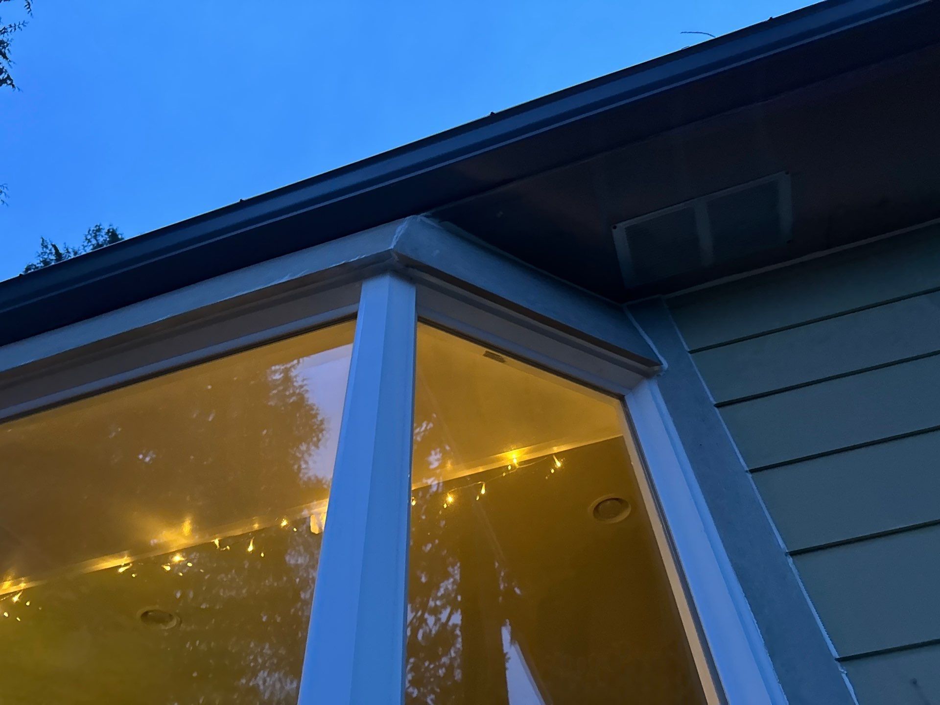 Blue house eaves and window frame at dusk, with warm yellow light reflecting in the glass.