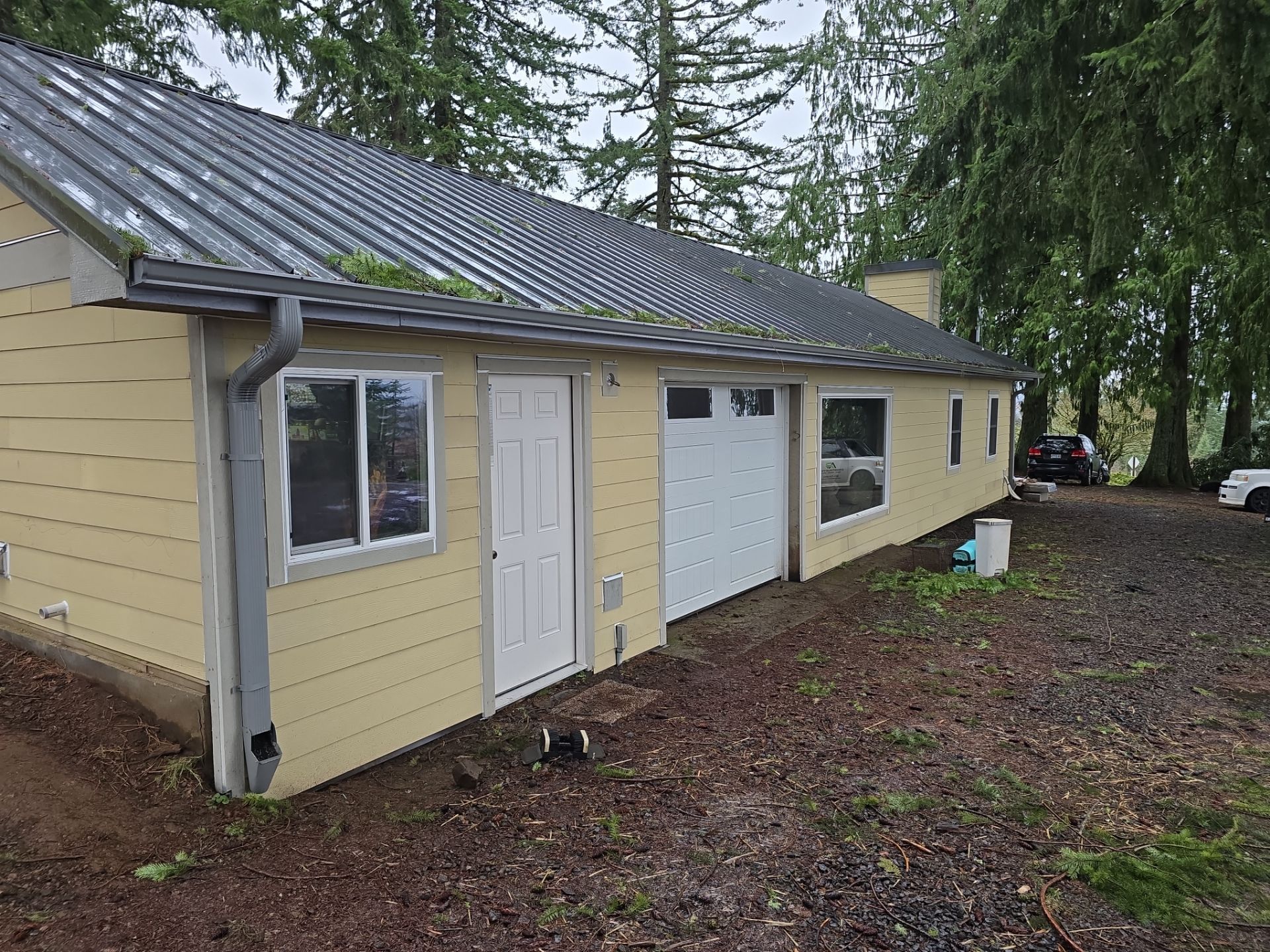 Yellow house with white doors and garage, wet yard, and tall trees nearby