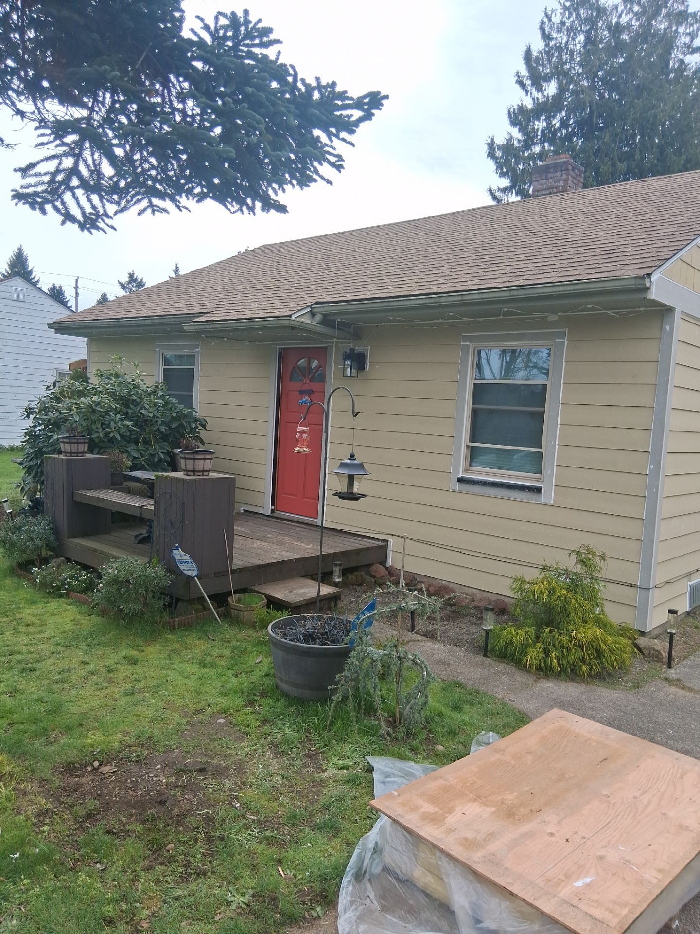 Small beige house with a red door, front porch, and grassy yard with plants and a wooden box.