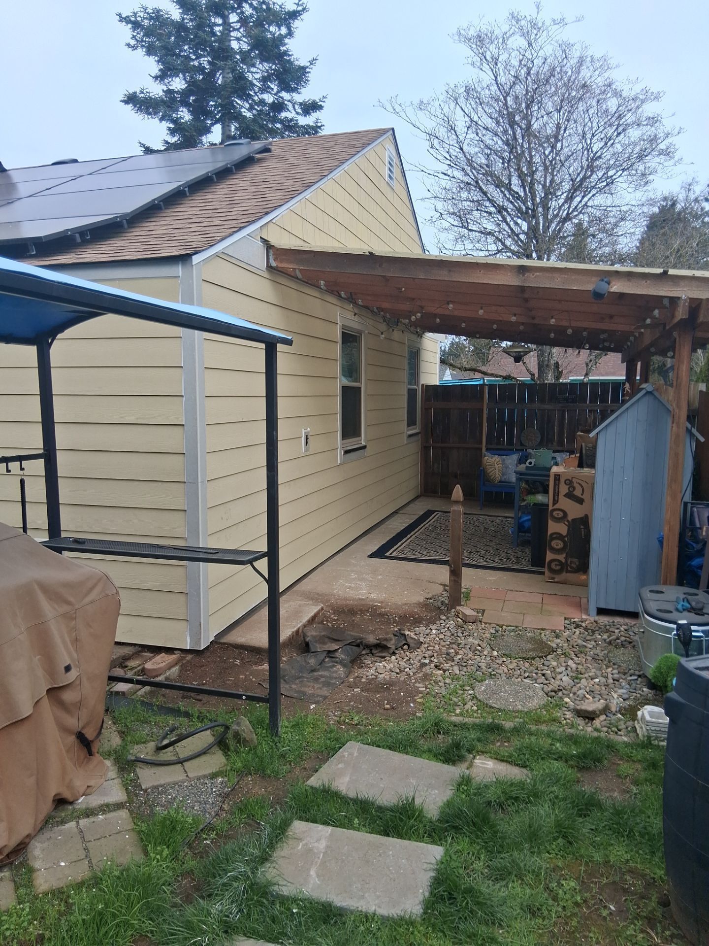 Backyard beside beige house with covered patio, fence, and stepping stones under overcast sky