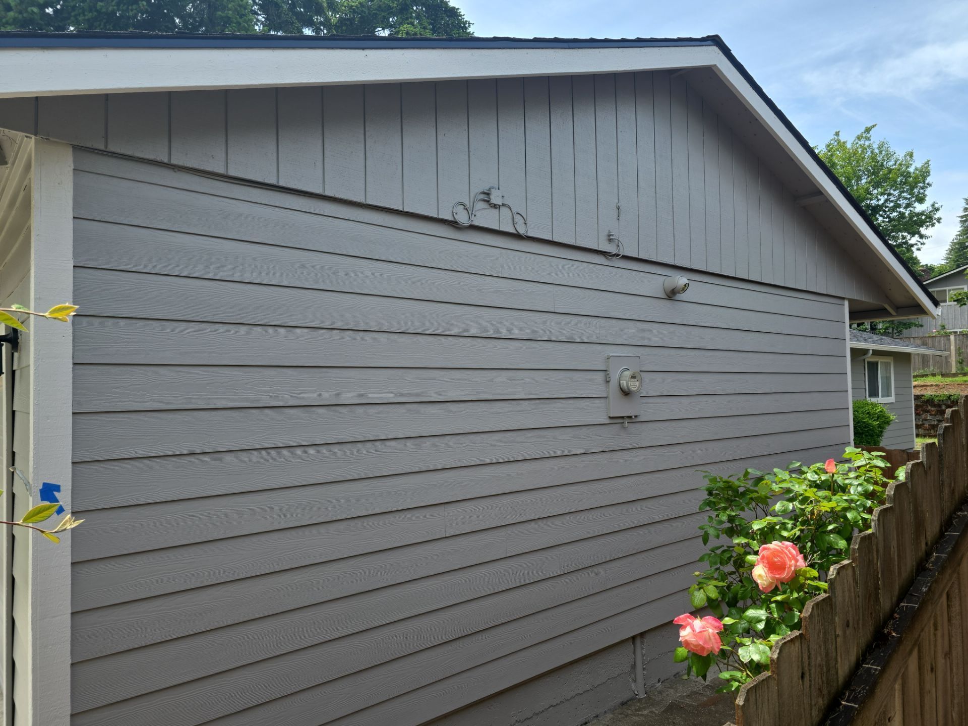 Gray house siding with a cracked upper wall, seen beside a fence and pink flowers in the foreground