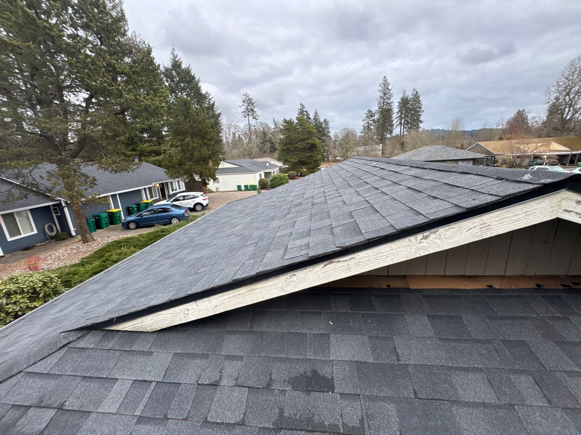 Collapsed roof ridge with missing shingles on a gray house in a suburban neighborhood under cloudy skies