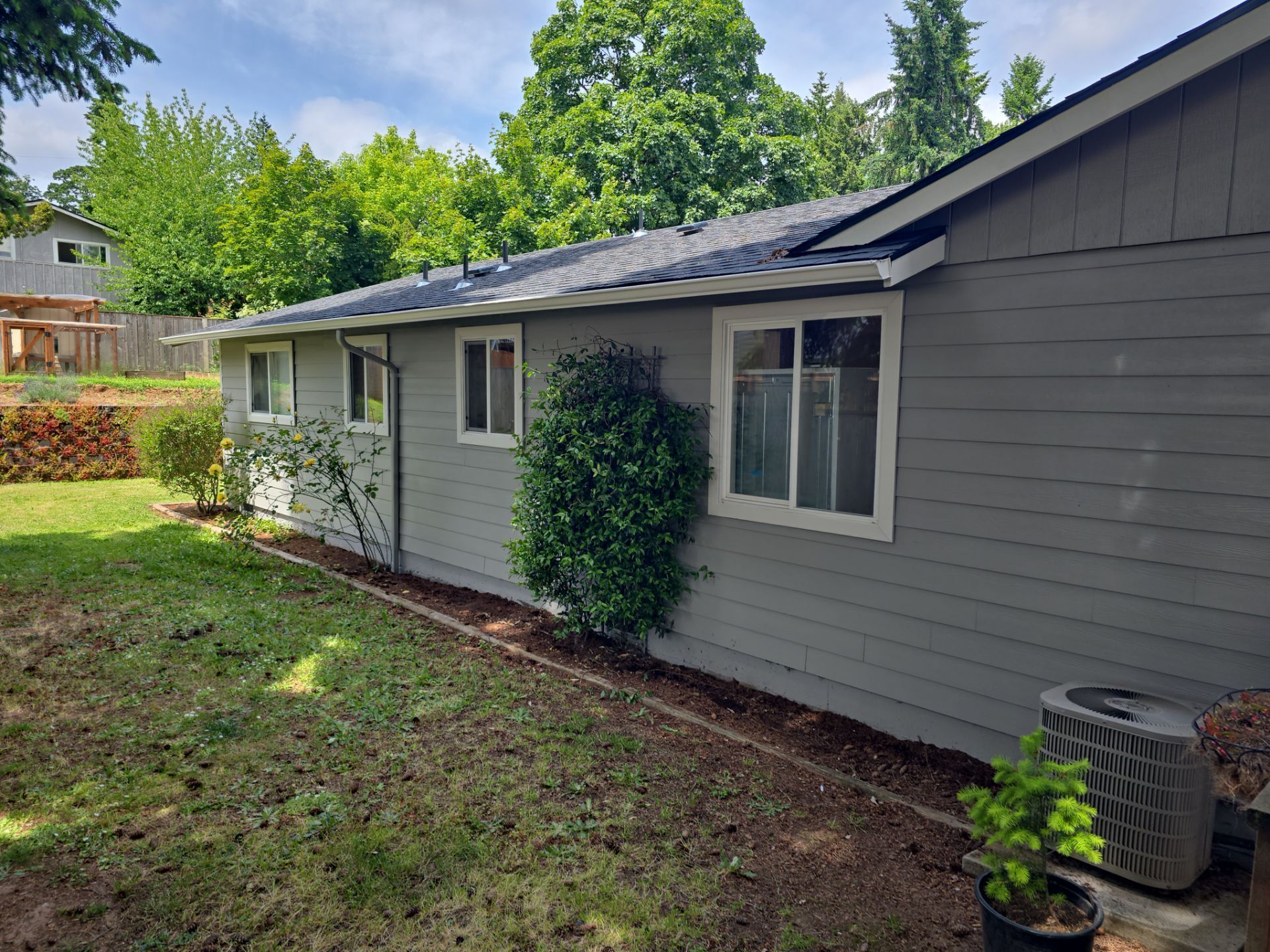 Gray house exterior with white-trimmed windows, dark roof, and small yard with trees and shrubs