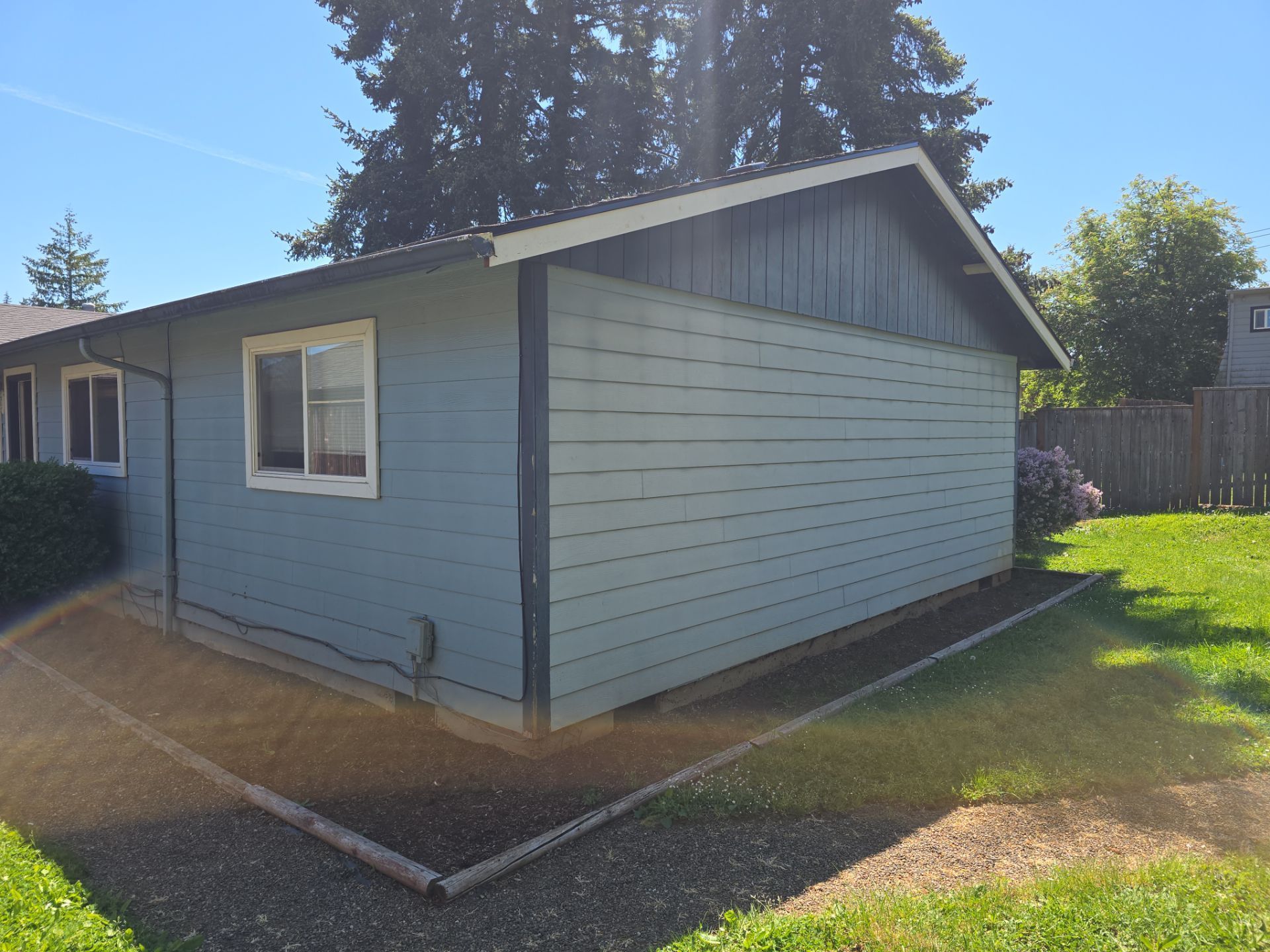 Blue-gray house exterior with white-trimmed windows and a sloped roof, beside a grassy yard and gravel path.