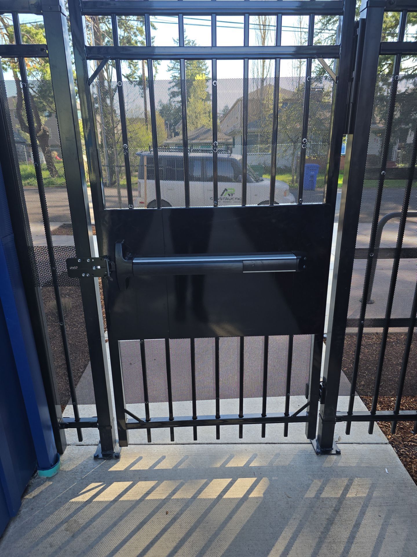 Black metal gate with a push bar, casting shadows on a concrete landing overlooking trees outside.