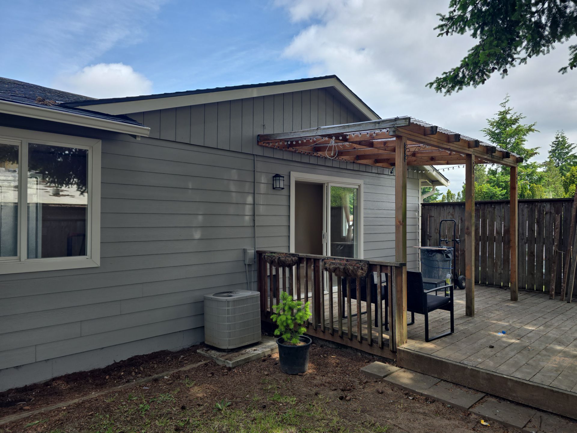 Backyard patio beside a gray house with a wooden pergola, railing, potted plant, and fenced yard.