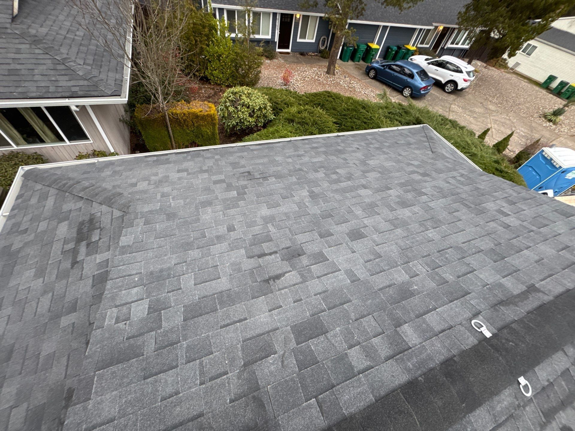 Gray shingled roof in a suburban neighborhood, viewed from above with houses and parked cars in the background