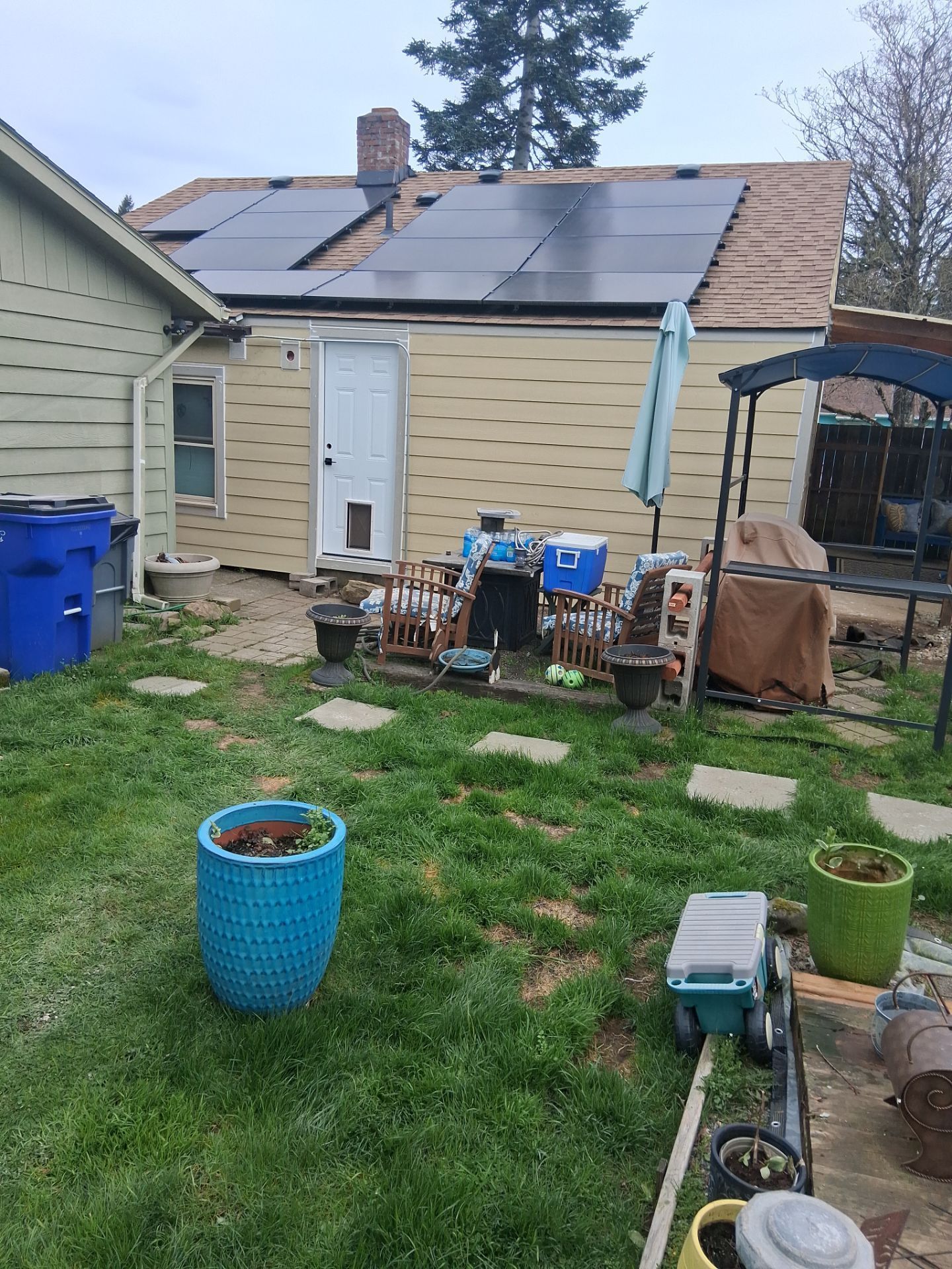 Backyard with a beige shed, solar panels on the roof, and scattered bins, planters, and garden tools.