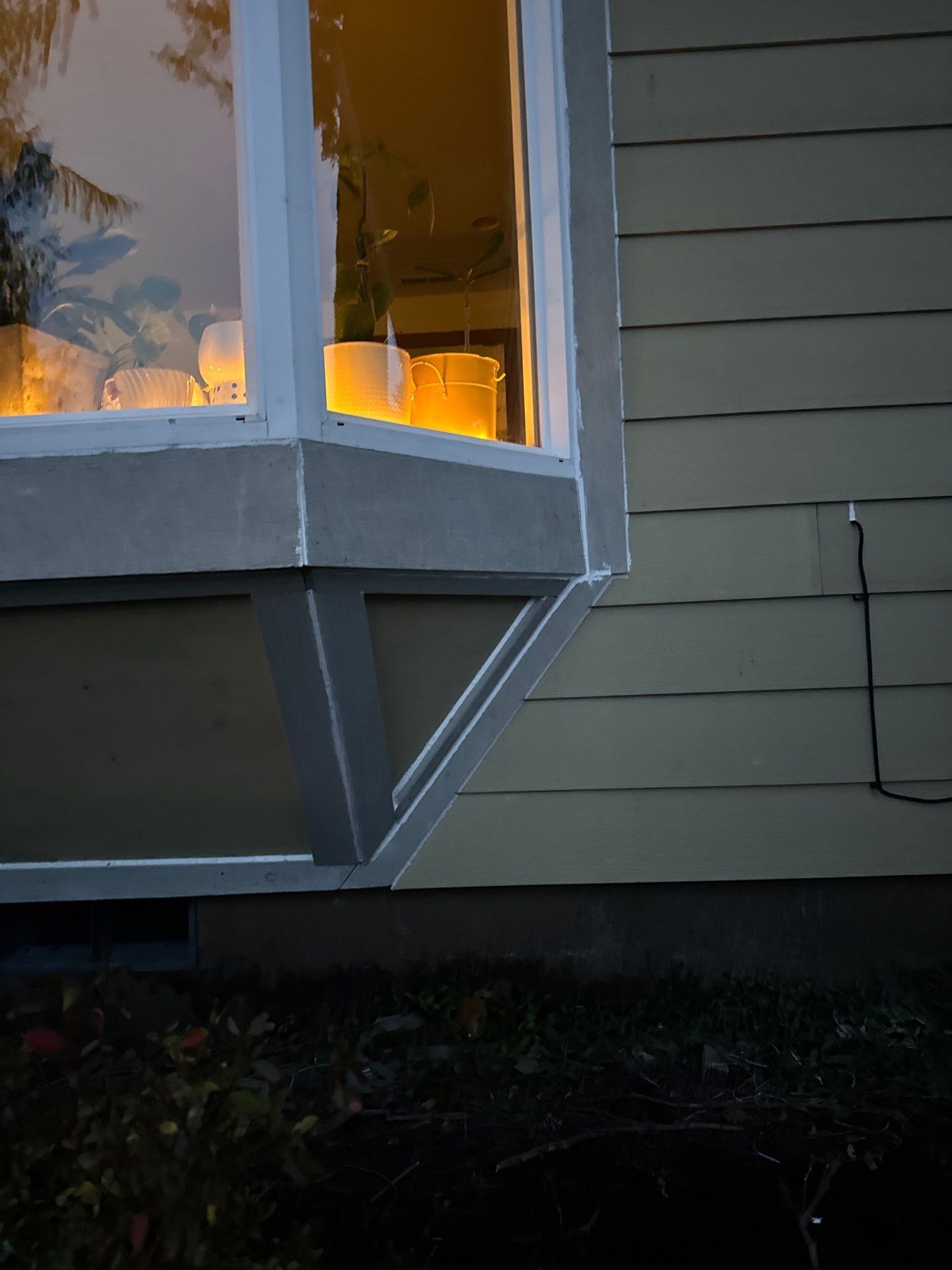 Lit bay window on a house exterior at dusk, with warm lights glowing inside and gray siding outside