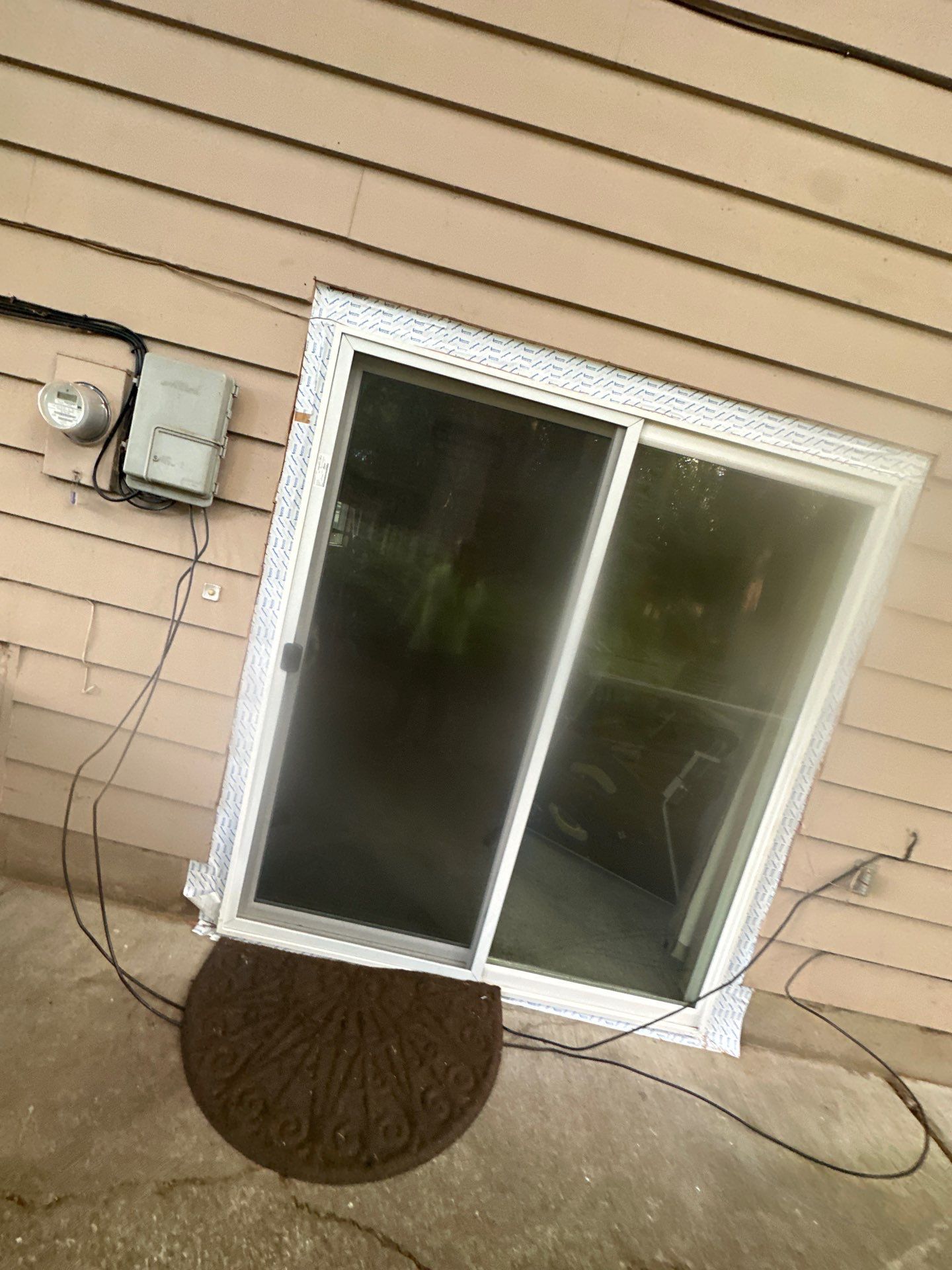 Sliding glass door in tan house siding, with utility boxes and cables on the left and a doormat below.