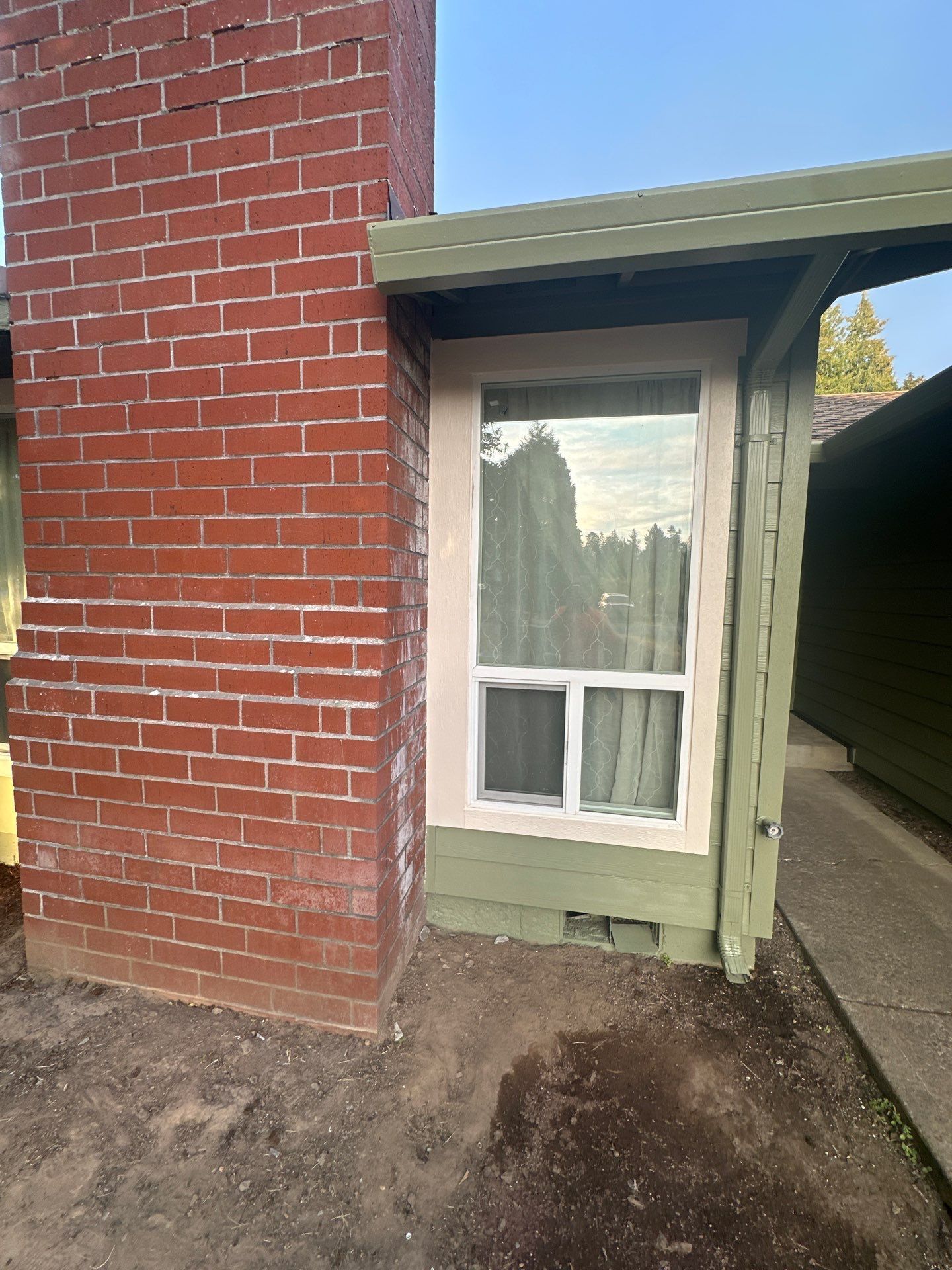 Red brick wall beside a small green-framed window under an awning, next to a narrow walkway.