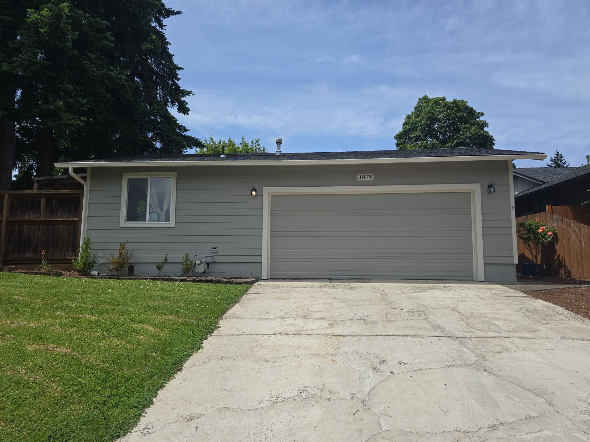 Single-story gray house with a two-car garage and concrete driveway, framed by lawn and trees.