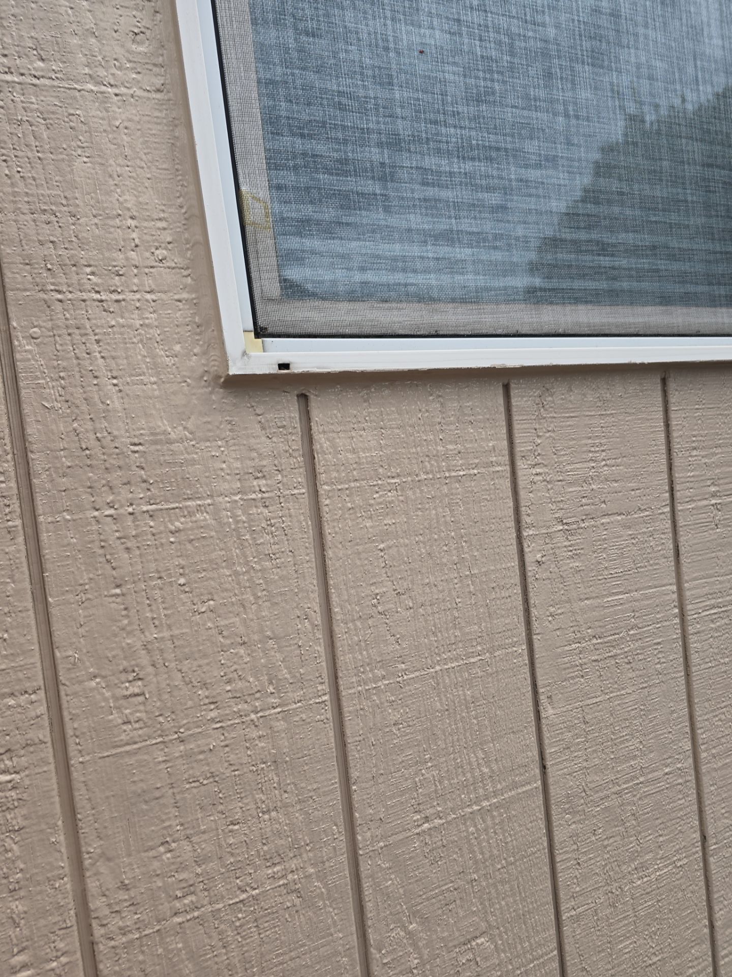 Beige exterior wall with vertical siding and a screened window in the upper right corner