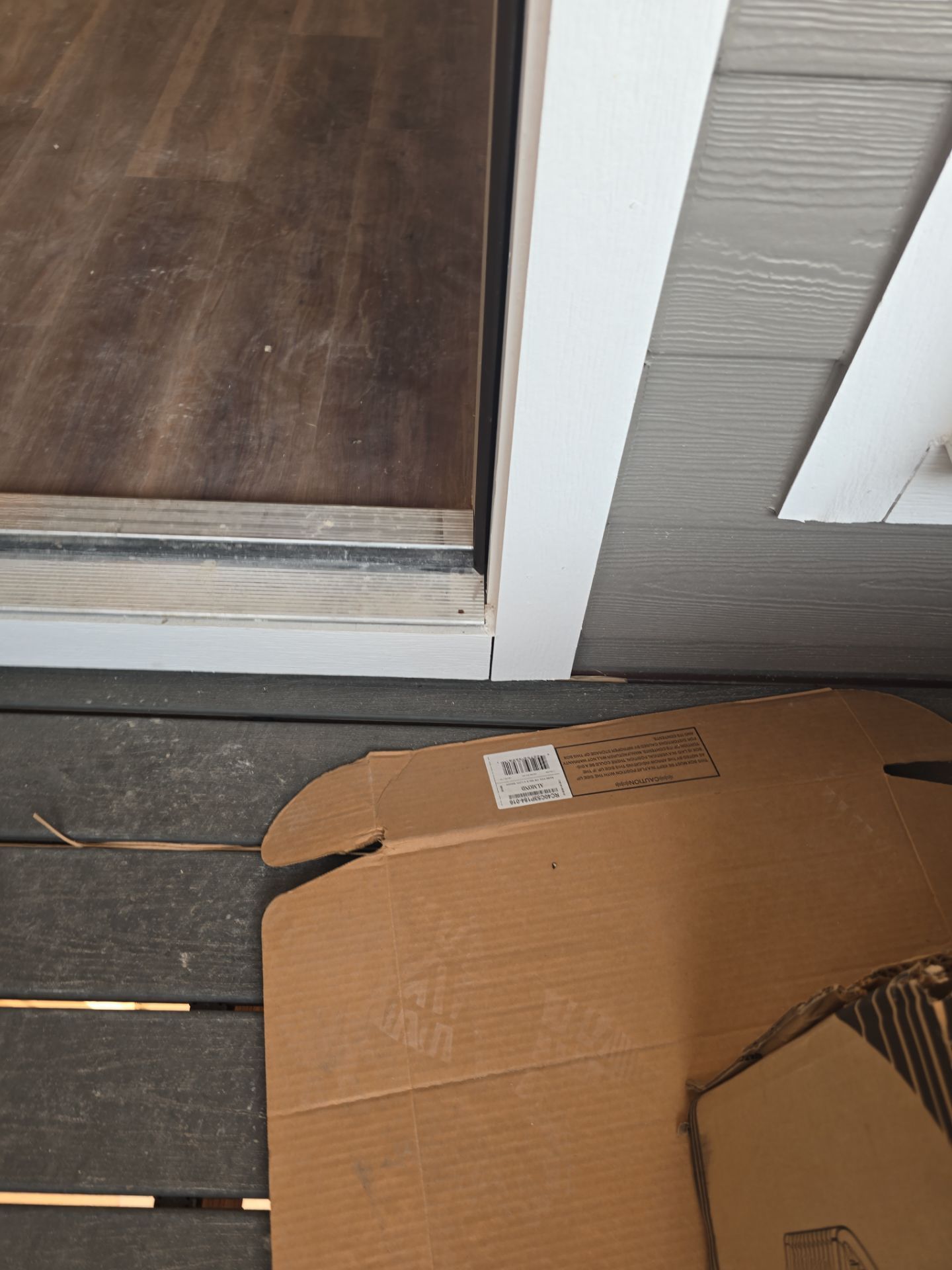 Cardboard box on a porch beside a closed white door and dark wood deck flooring.