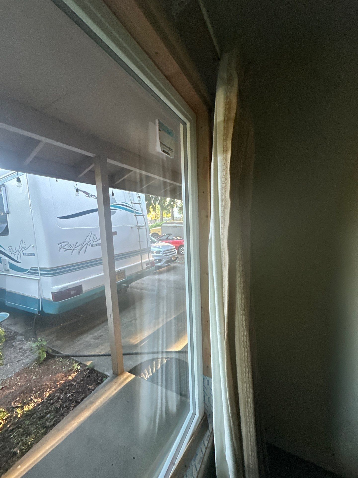 View through a window with curtains, showing a rainy street, parked RV, and a car outside.
