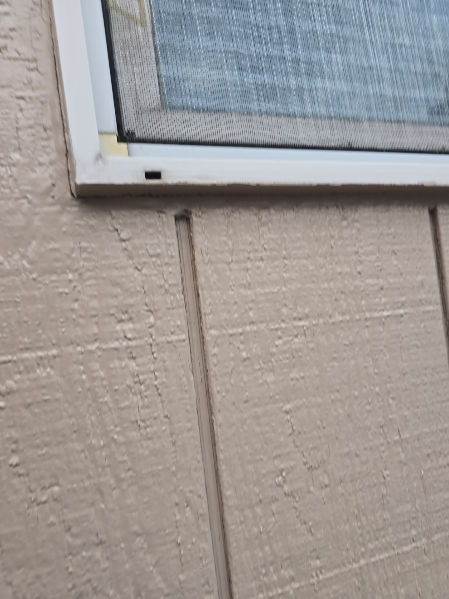 Close-up of a window frame with a cracked beige wall below it and a vertical seam in the siding.