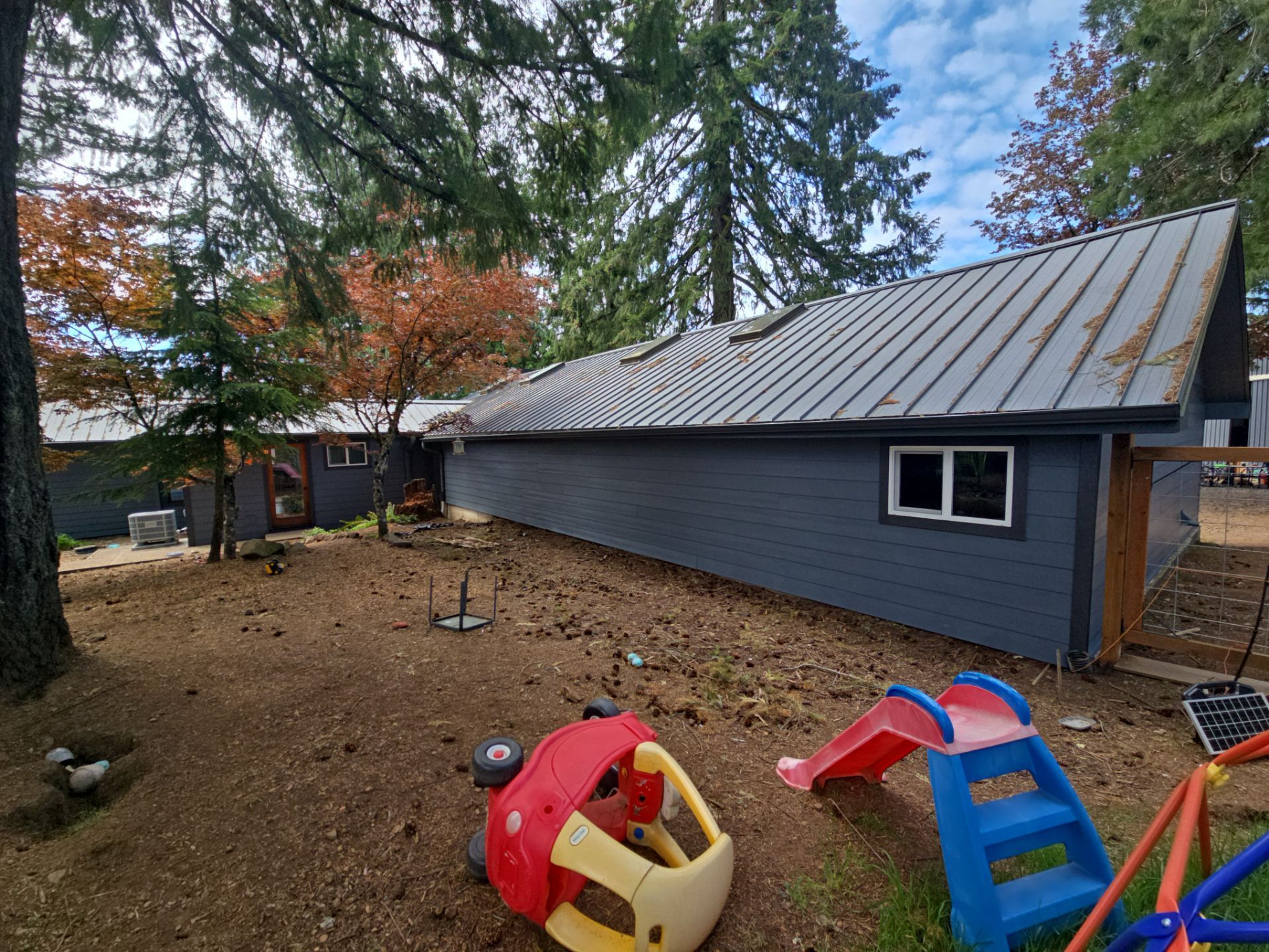 Small yard with play equipment beside a gray house and tall trees in autumn.