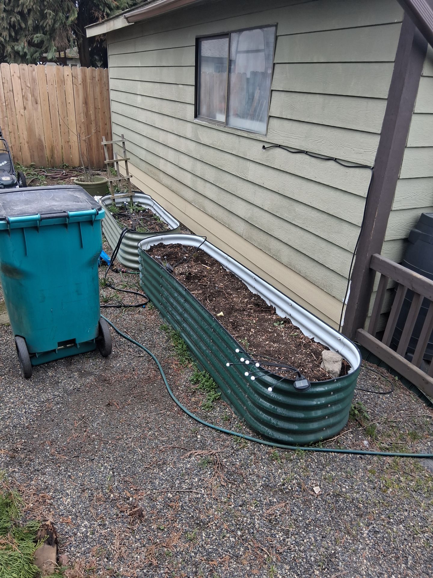 A blue trash bin beside a green planter and a house wall along a gravel path.
