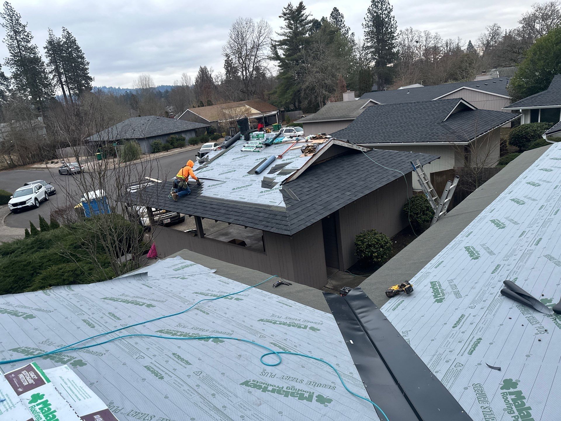 Rooftops under repair with workers, exposed underlayment, and trees in a suburban neighborhood