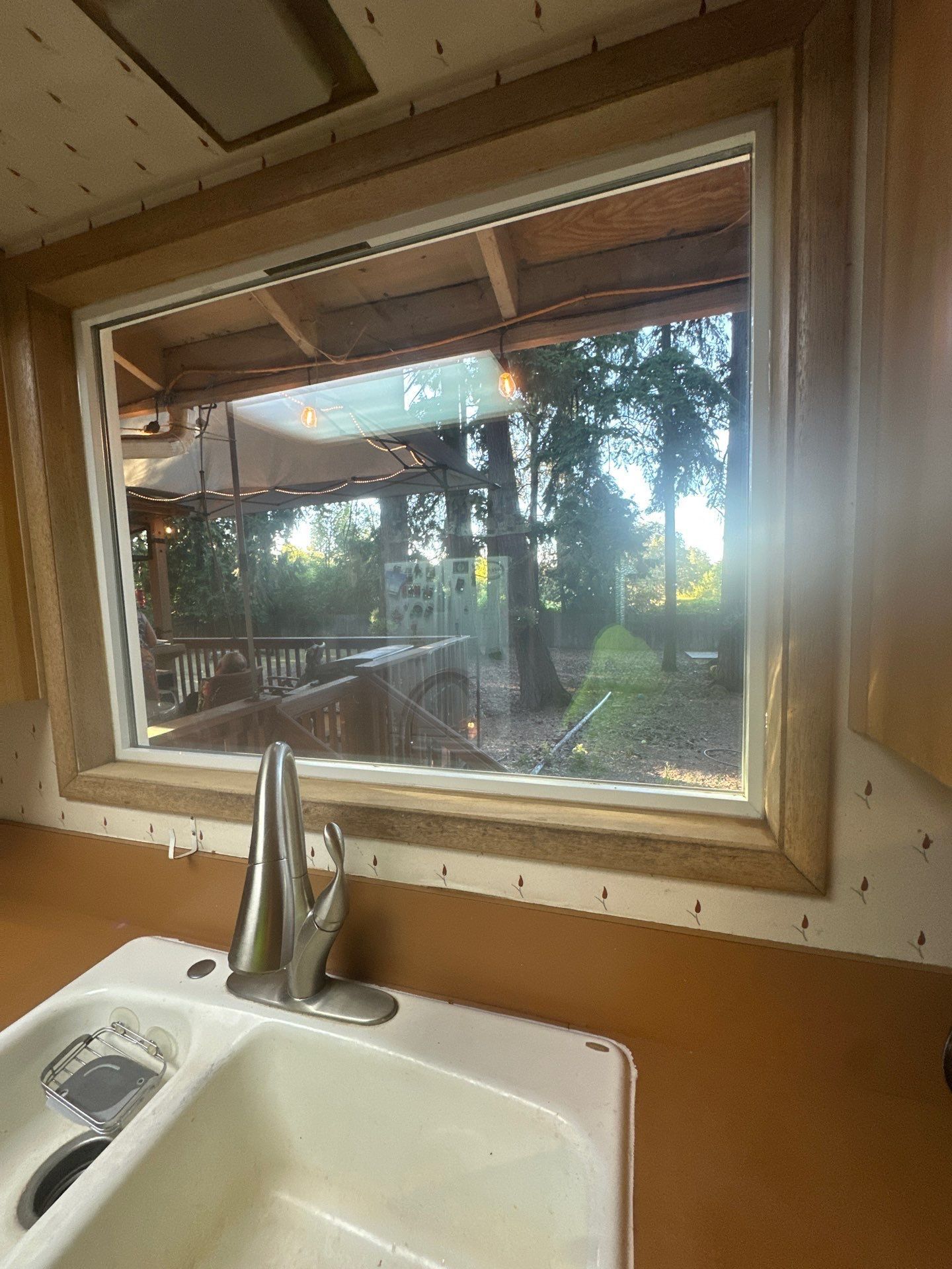 Kitchen sink beneath a window overlooking a sunlit backyard with trees and a deck