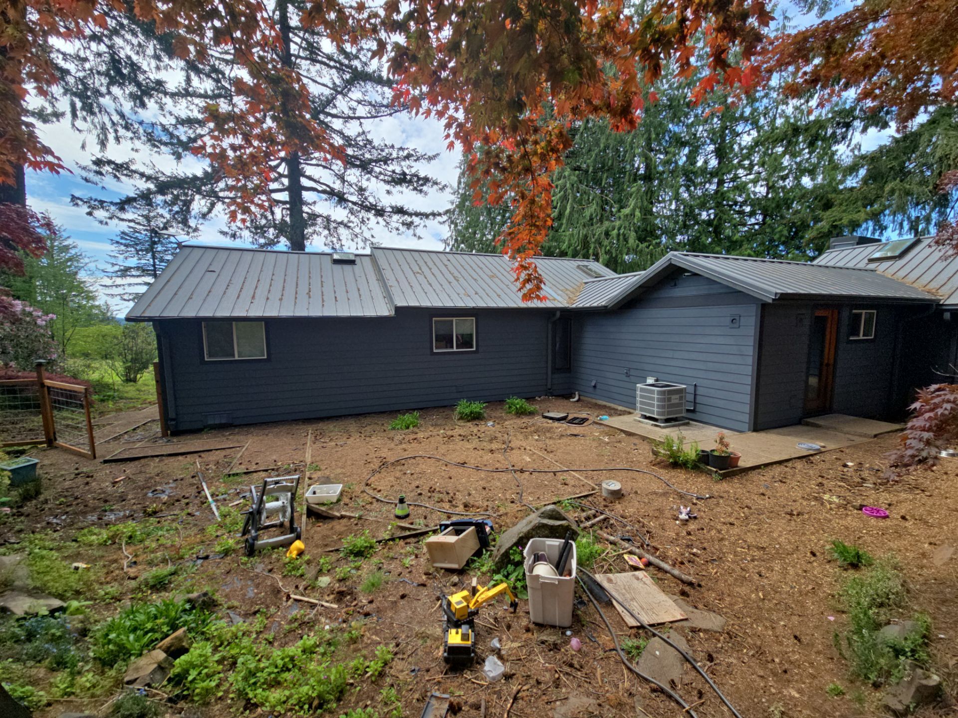 Backyard view of a dark house with gray addition, surrounded by trees and a bare garden bed