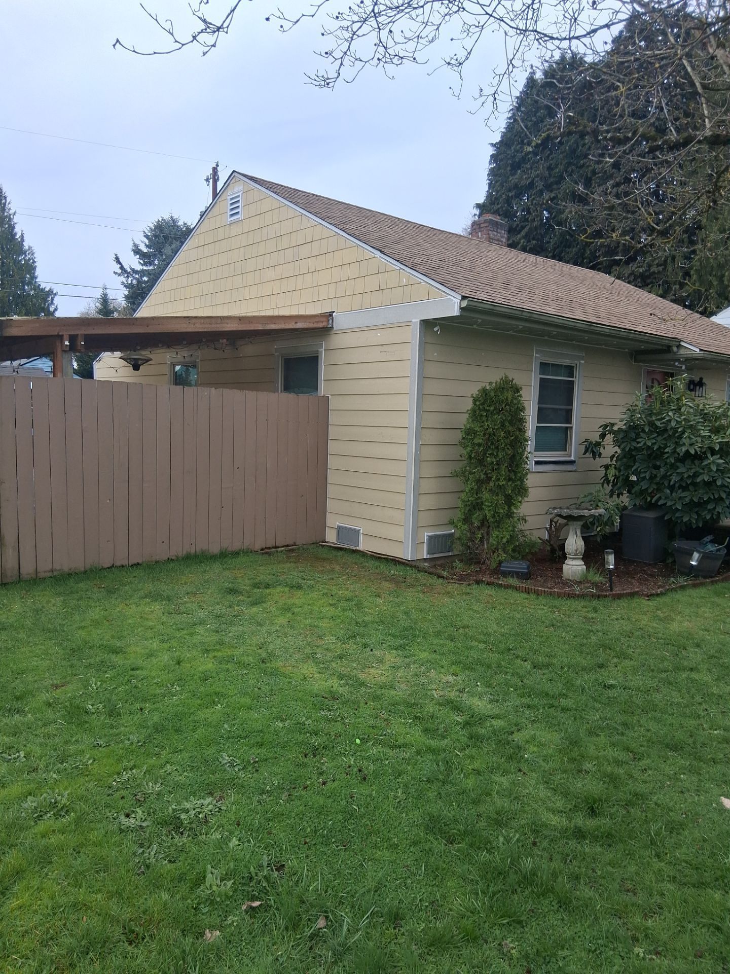 Small beige house with a tan roof, fenced yard, green lawn, and tall tree at the right side.