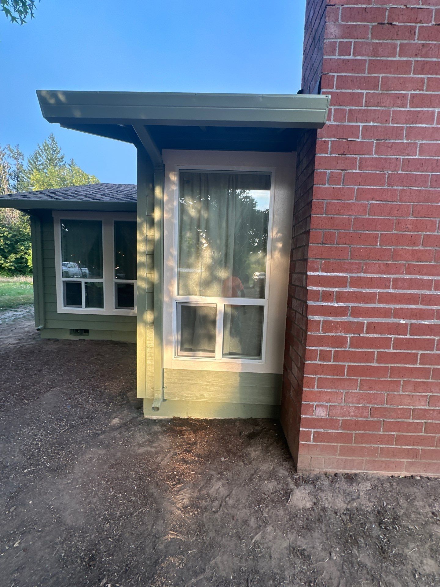 Small green house entrance with a covered porch, window, and red brick wall beside a gravel path.
