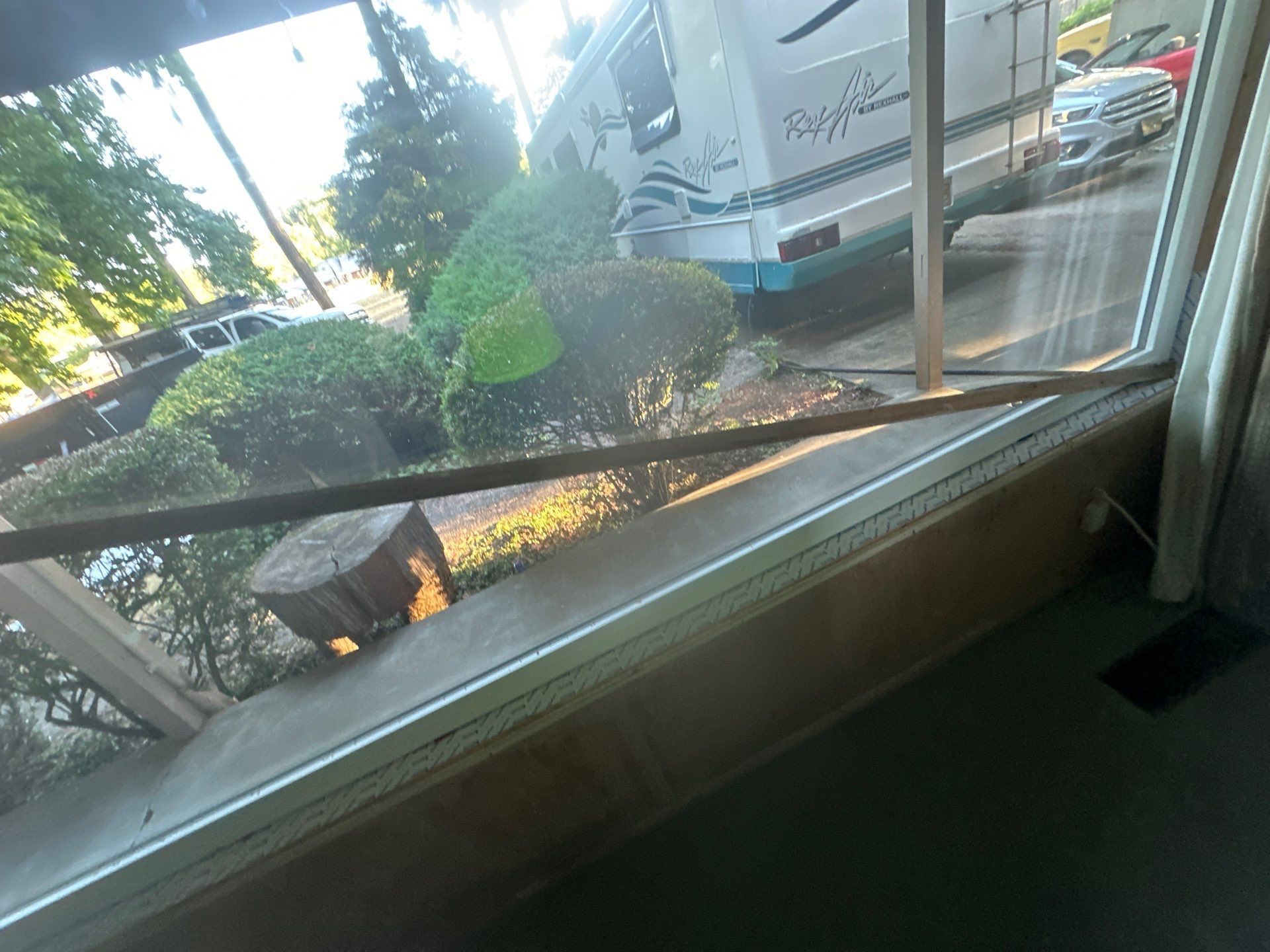 View through a window of a sidewalk and parking area with trees, bushes, and a truck.