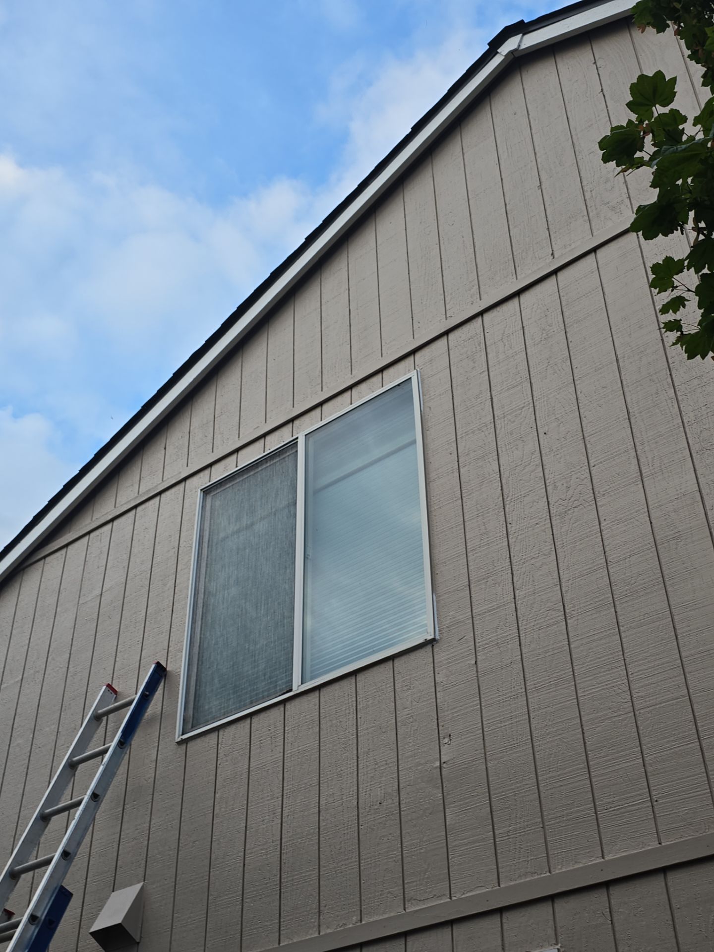 Side of a beige house with a ladder leaning against it and a window near the roofline under a blue sky.